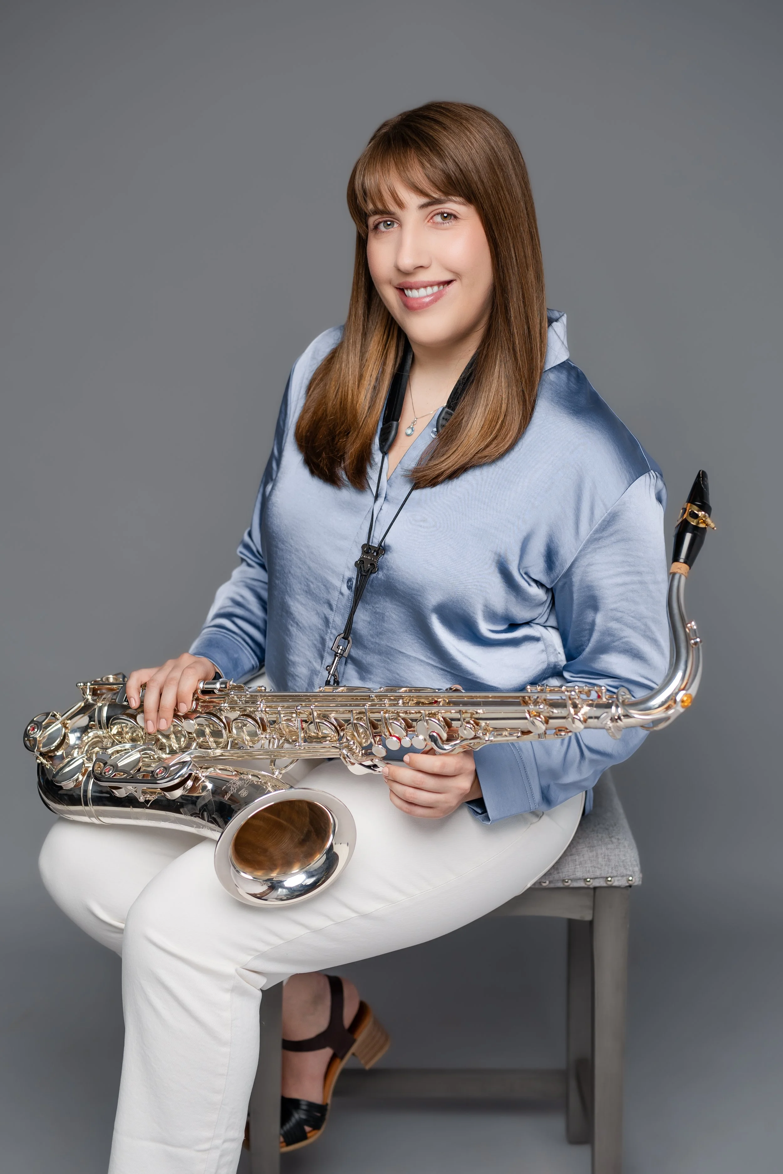 A woman with long brown hair and light skin, smiling, sitting on a gray stool, holding a silver saxophone, wearing a blue satin blouse with white pants, in front of a plain gray background.