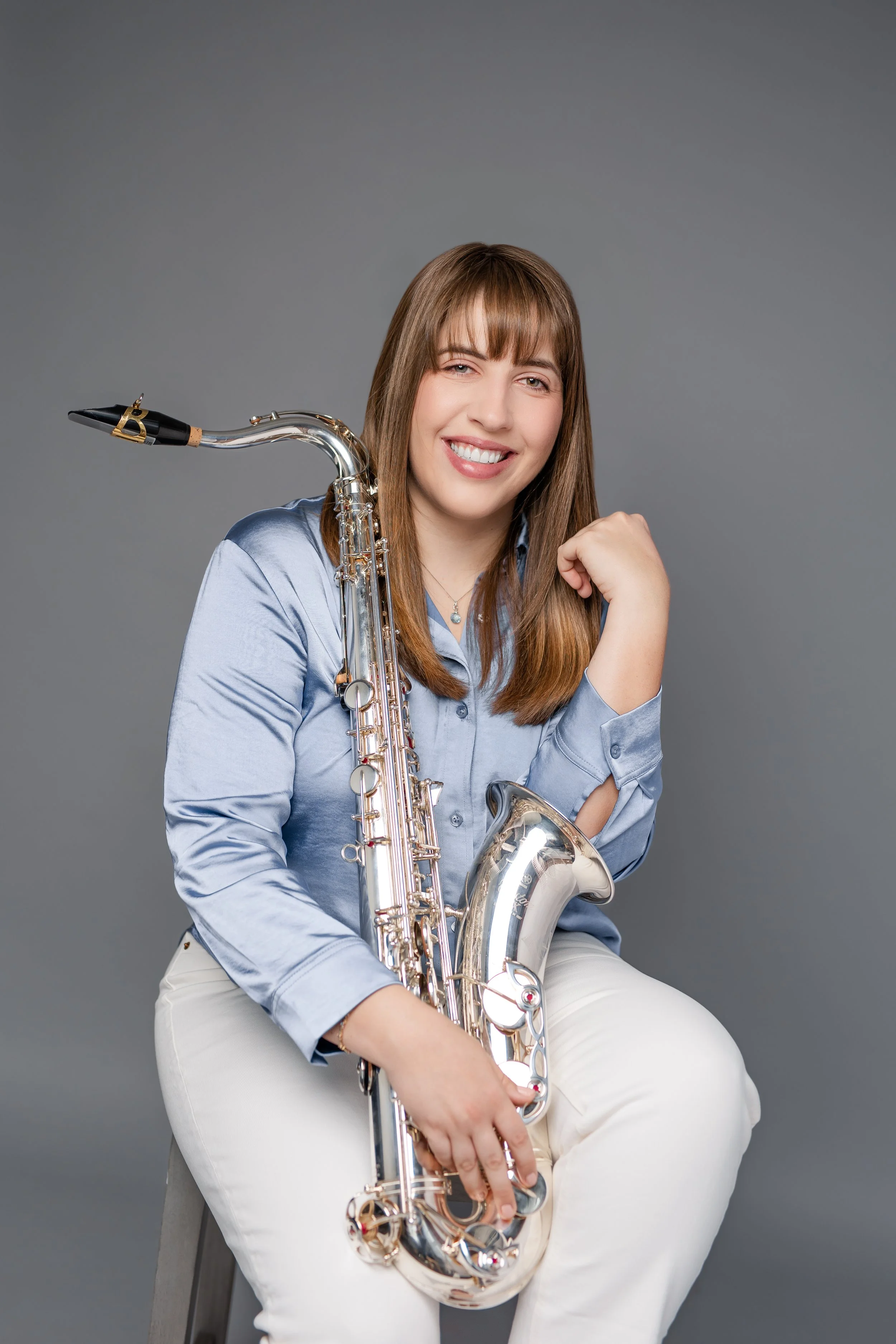 A young woman with long brown hair and bangs smiling and holding a saxophone, wearing a light blue button-up shirt and white pants, against a plain gray background.