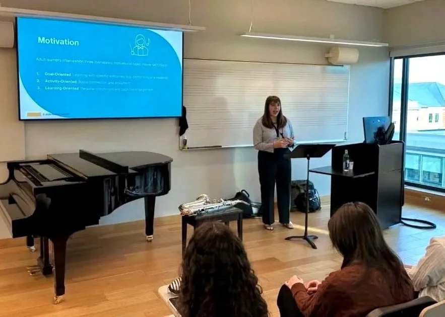 A woman giving a lecture on motivation in a classroom with a piano, a large window, and students seated facing her.
