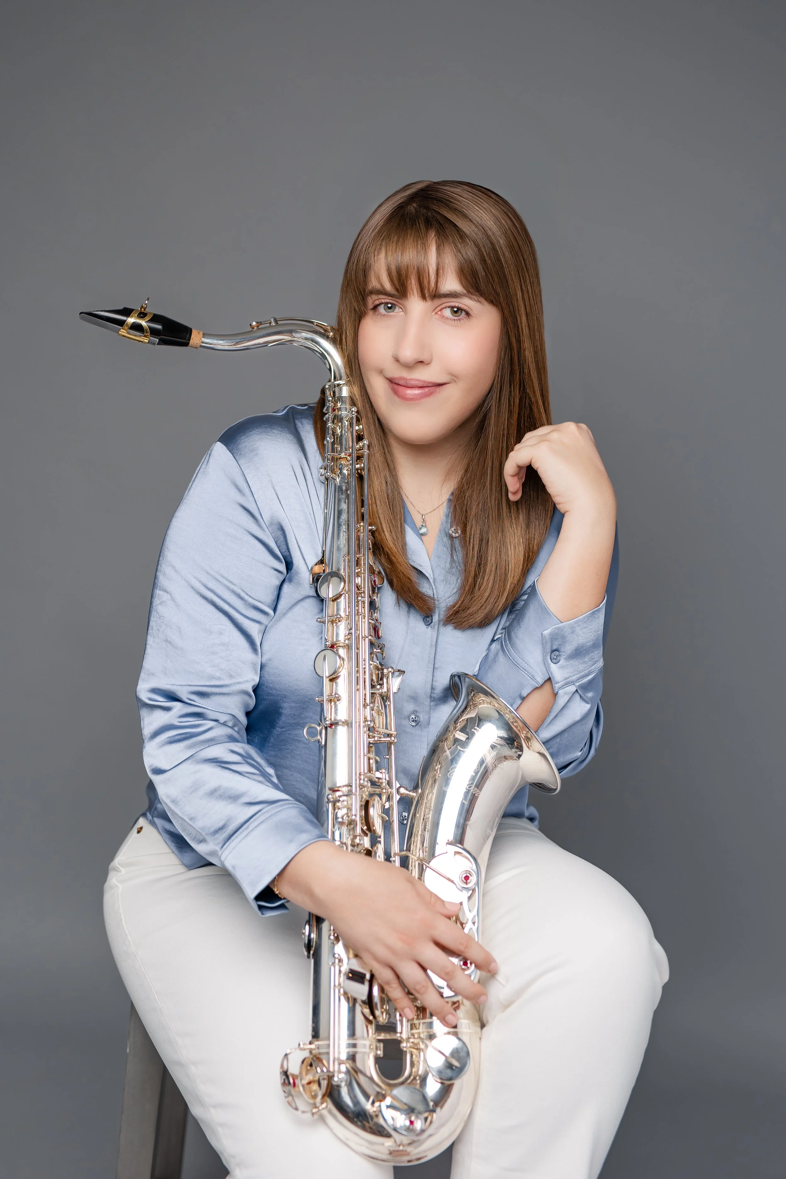 A young woman with brown hair and bangs, wearing a light blue shirt and white pants, sitting with a silver saxophone against a gray background, smiling at the camera.