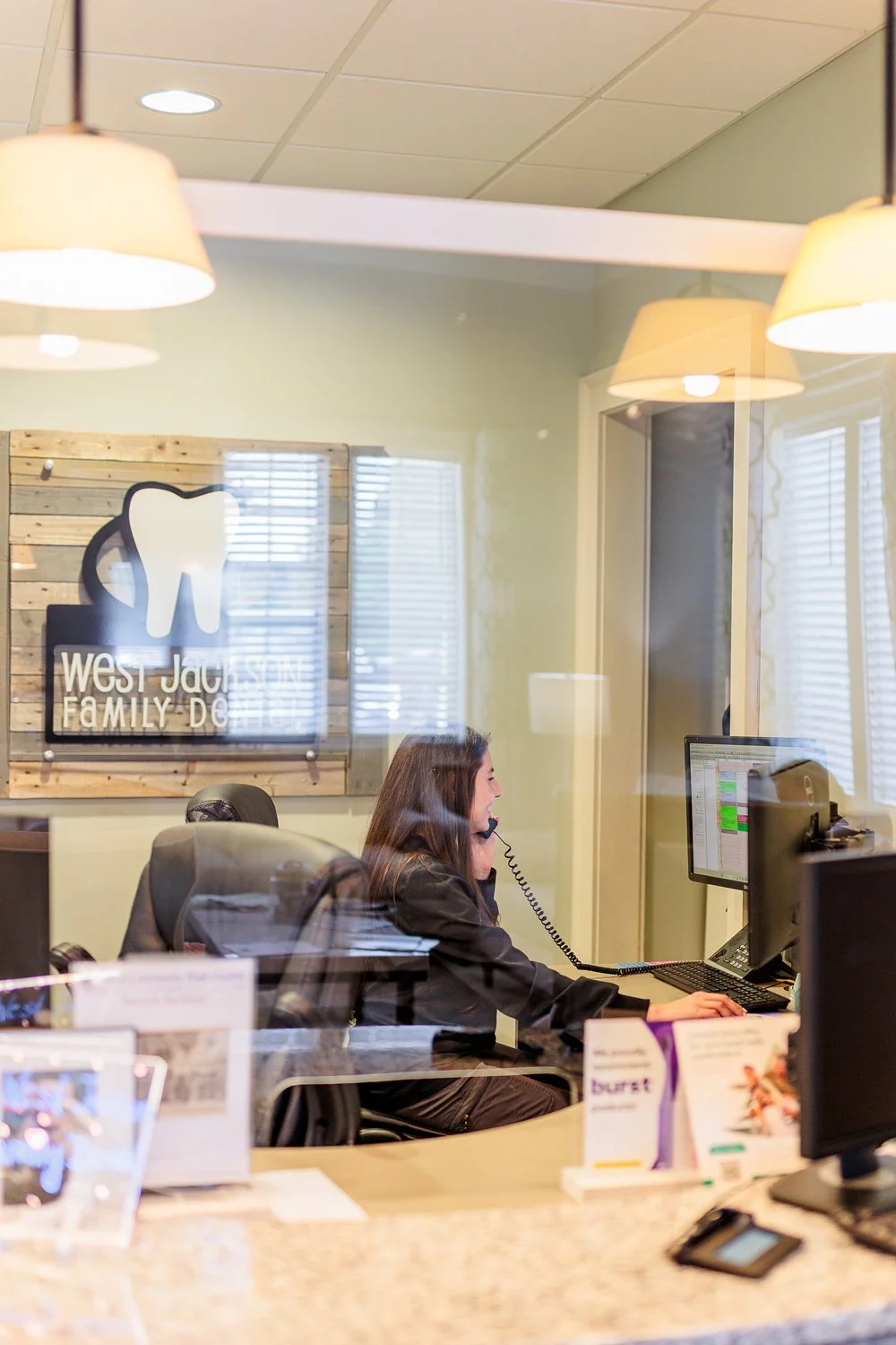 Office reception area with woman on phone at desk, West Jackson Family Dental sign on wooden wall, and office equipment visible through glass window.
