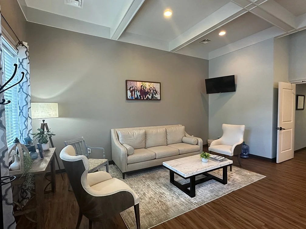 Living room with a white sofa, armchairs, a marble-topped coffee table, flat-screen TV, framed photo, and a window with patterned curtains.