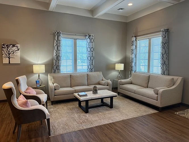 Living room with two beige sofas, two white armchairs with pink pillows, two lamps on side tables, a coffee table, a large area rug, and three windows with patterned curtains.