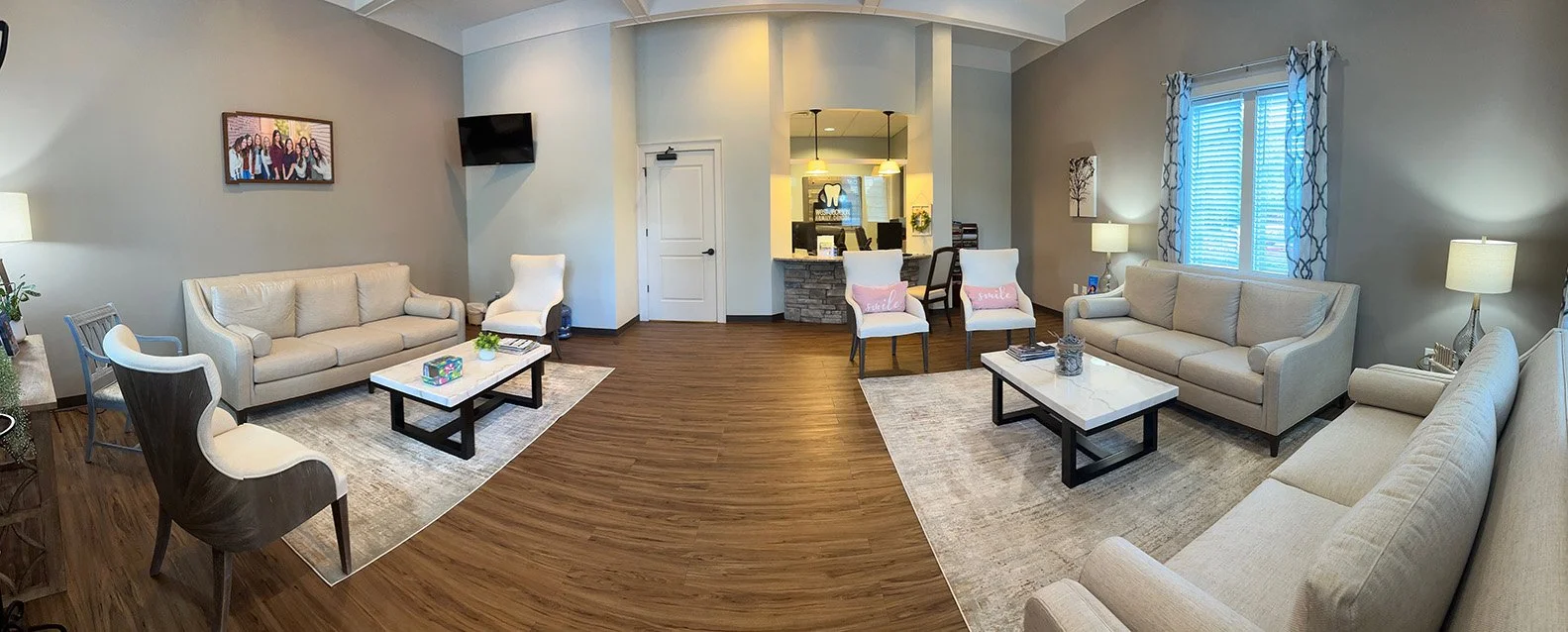 A spacious living room with beige sofas, armchairs, coffee tables, a wall-mounted TV, framed pictures, and large windows with patterned curtains.