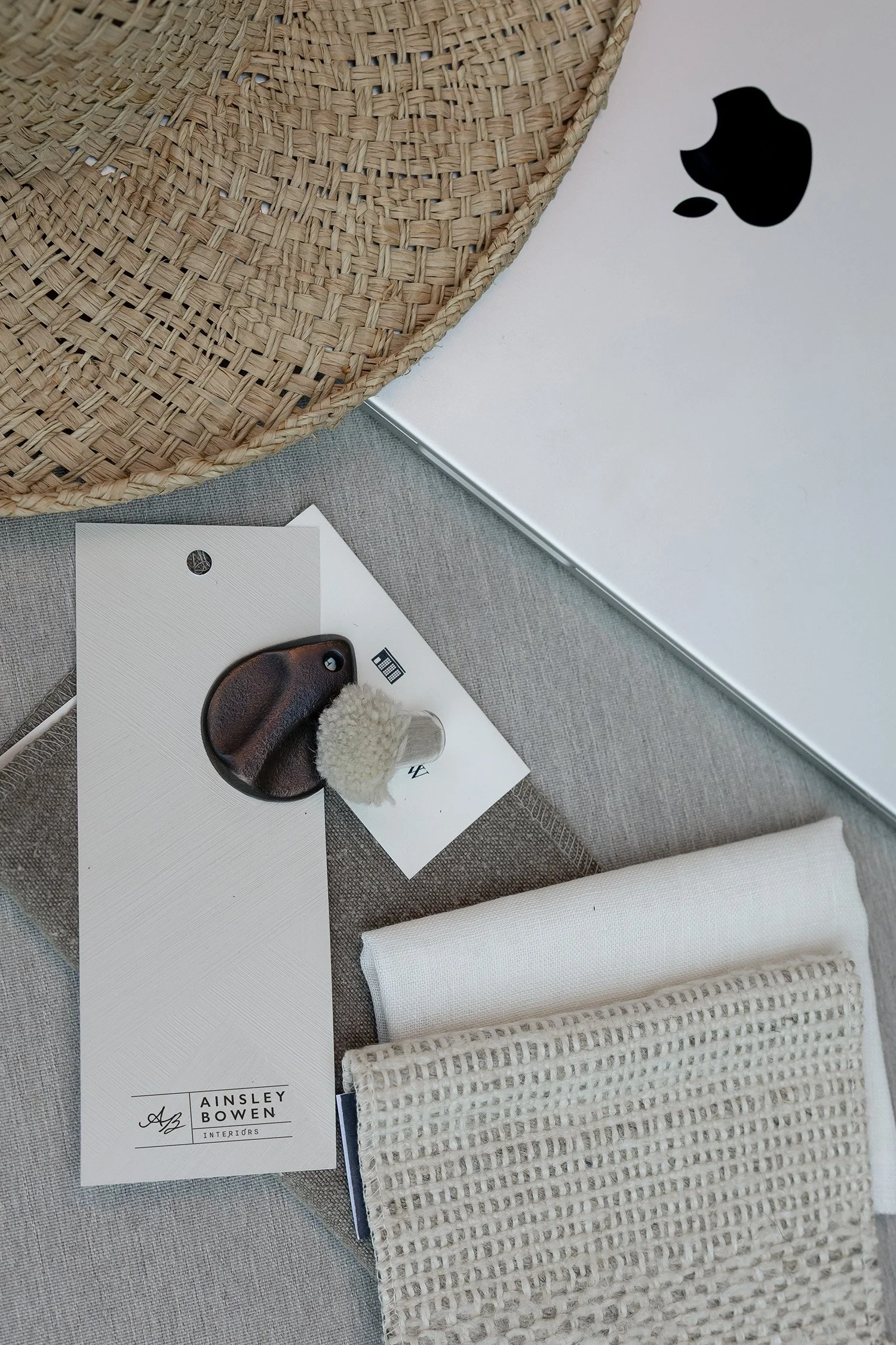 A silver laptop with a black apple logo, a woven wicker tray, fabric swatches, and an interior design mockup with a lamp and sample materials.