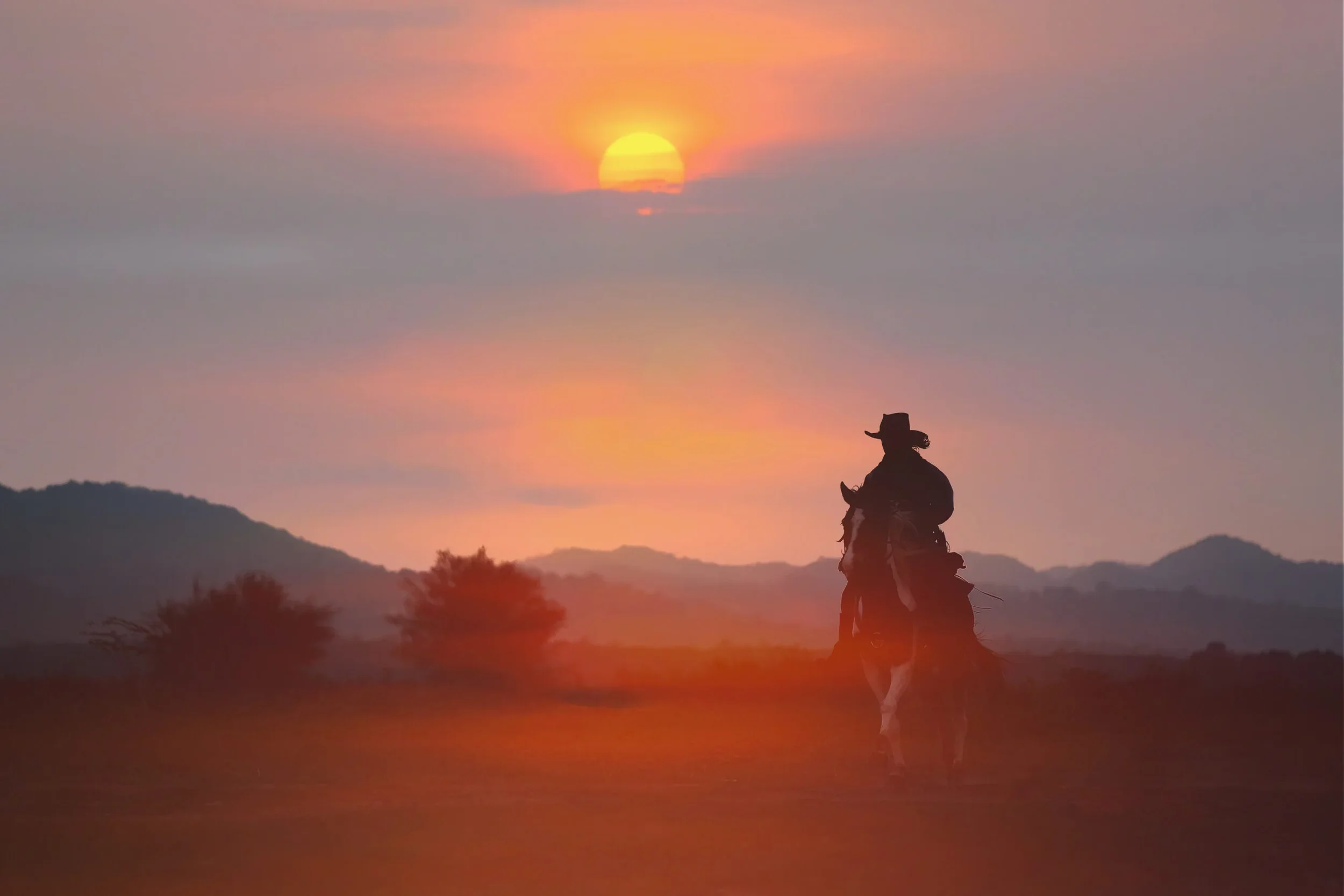 Silhouette of a cowboy riding a horse at sunset with mountains in the background.