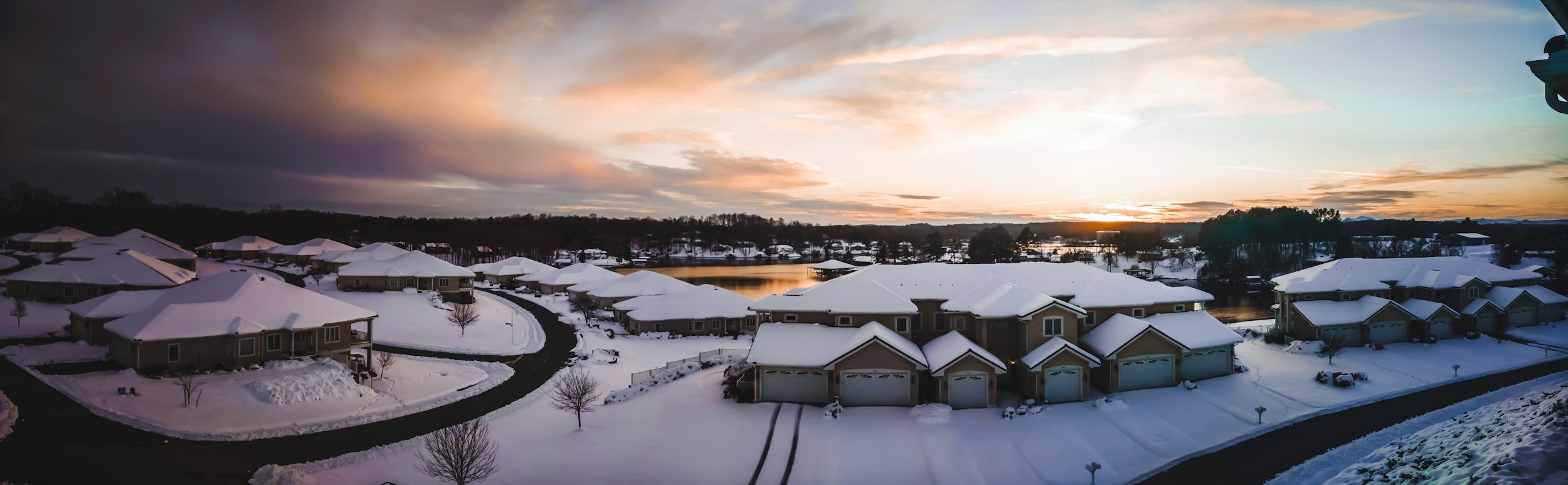 Aerial view of a snowy suburban neighborhood at sunset, showing houses with snow-covered roofs, driveways, and leafless trees, beside a body of water.