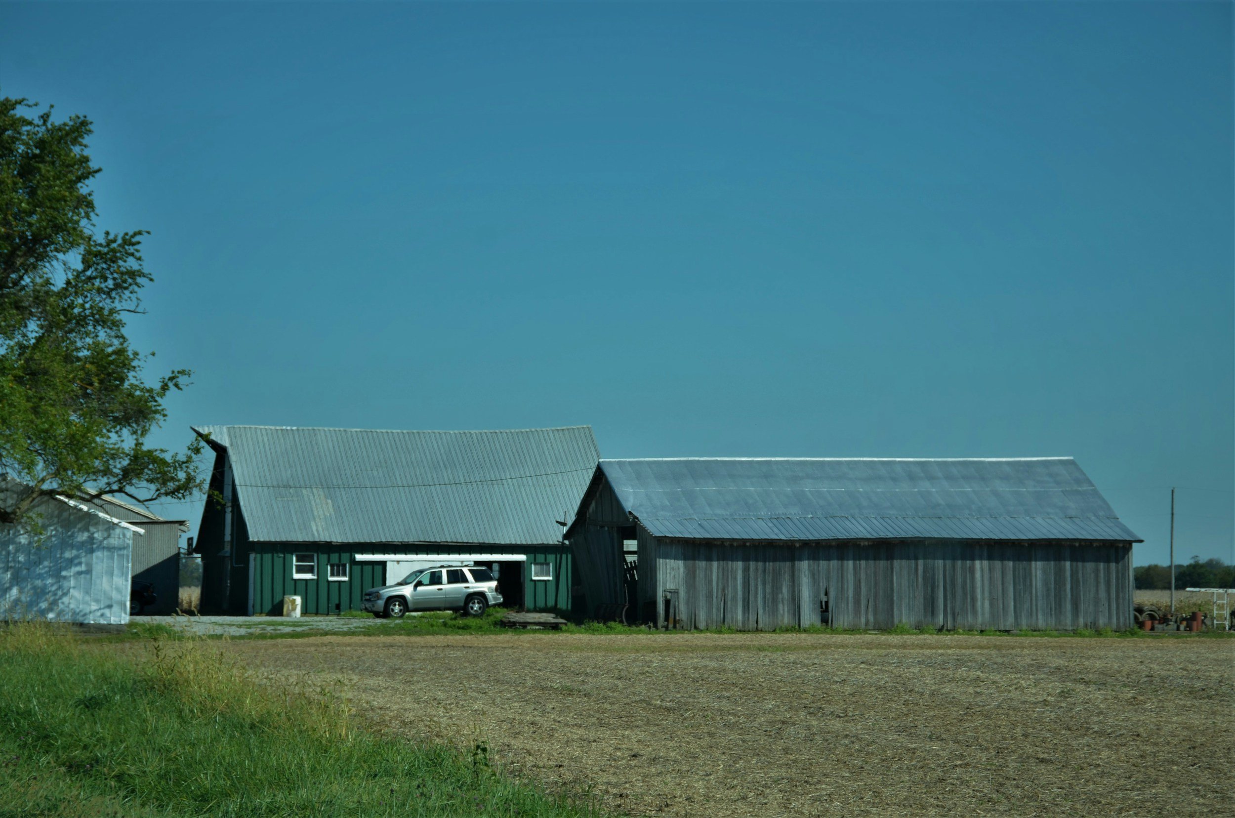 A rural farm scene with a green barn, a weathered gray barn, and a white vehicle parked in front of the barns, surrounded by open land and a blue sky.