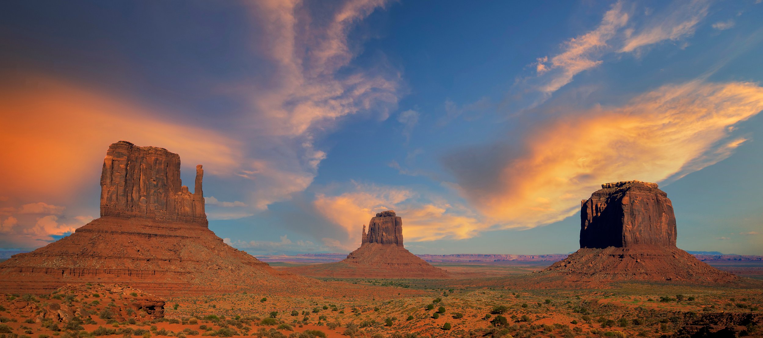 Three large sandstone buttes against a colorful sunset sky with clouds over a desert landscape