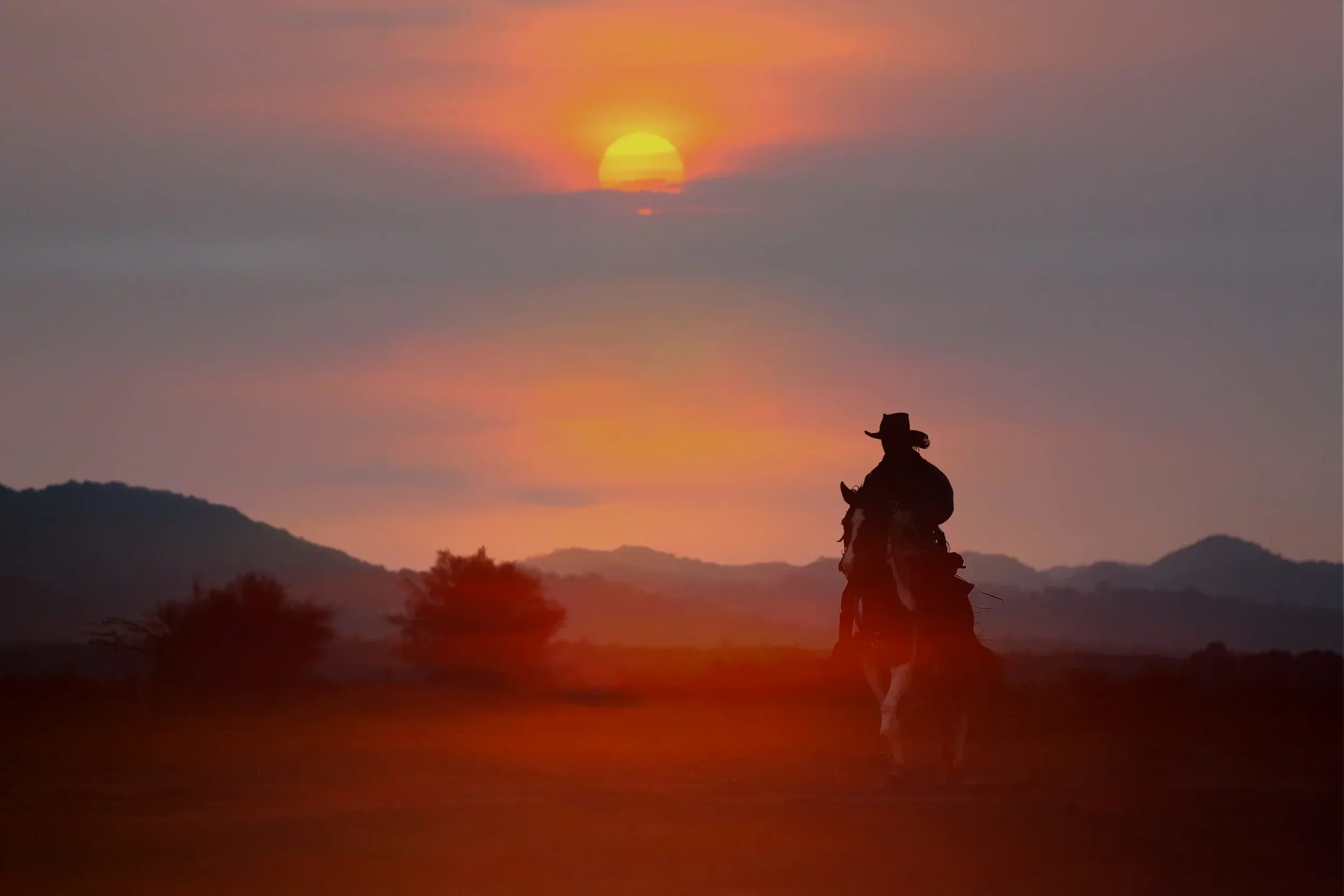 A silhouette of a cowboy riding a horse at sunset with a mountain range in the background and the sun partially covered by clouds.