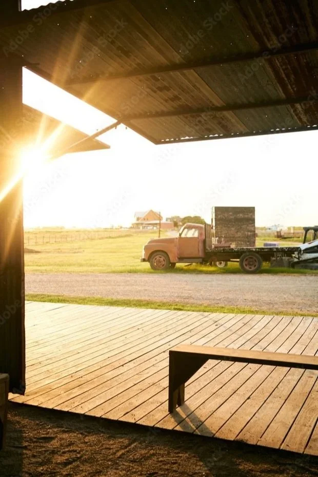 A vintage rusty pickup truck parked on a gravel area outside a barn or shed, with a grassy field and a small building with a red roof in the background during sunset, viewed from inside a wooden structure.