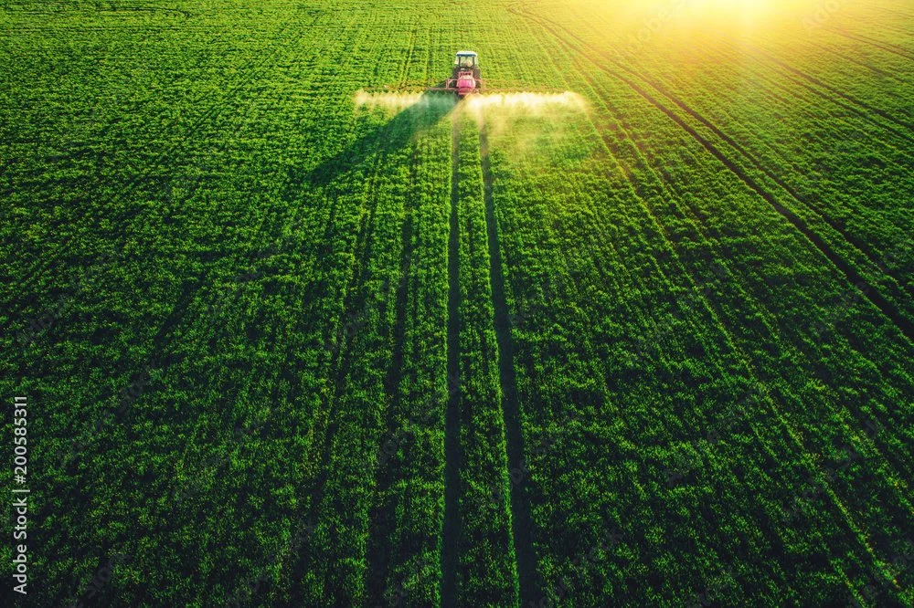 A tractor spray-paints a vast green crop field during sunset or sunrise.