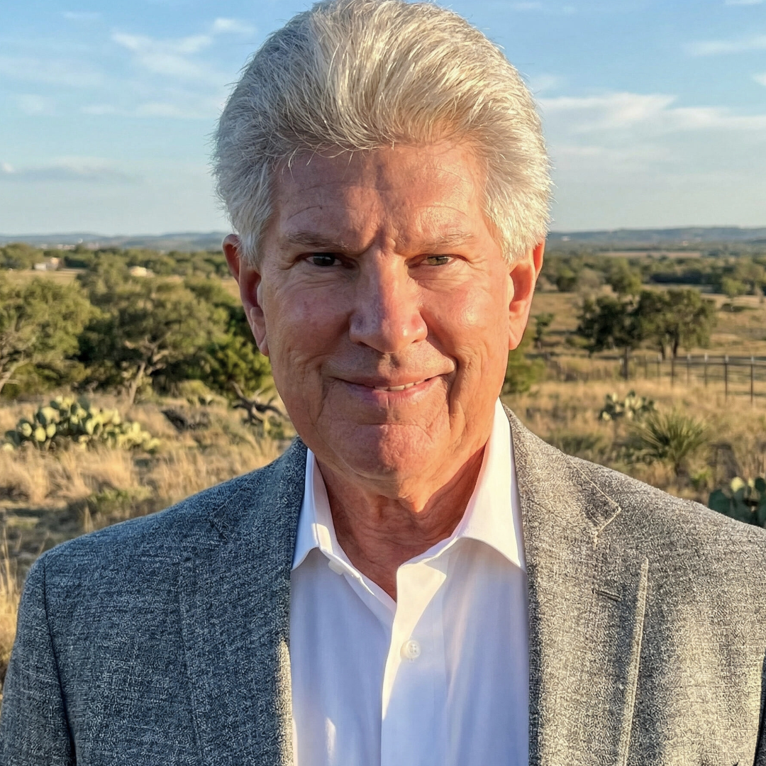 A smiling older man with white hair wearing a gray blazer and white shirt outdoors in a rural landscape with trees, cacti, and open fields.