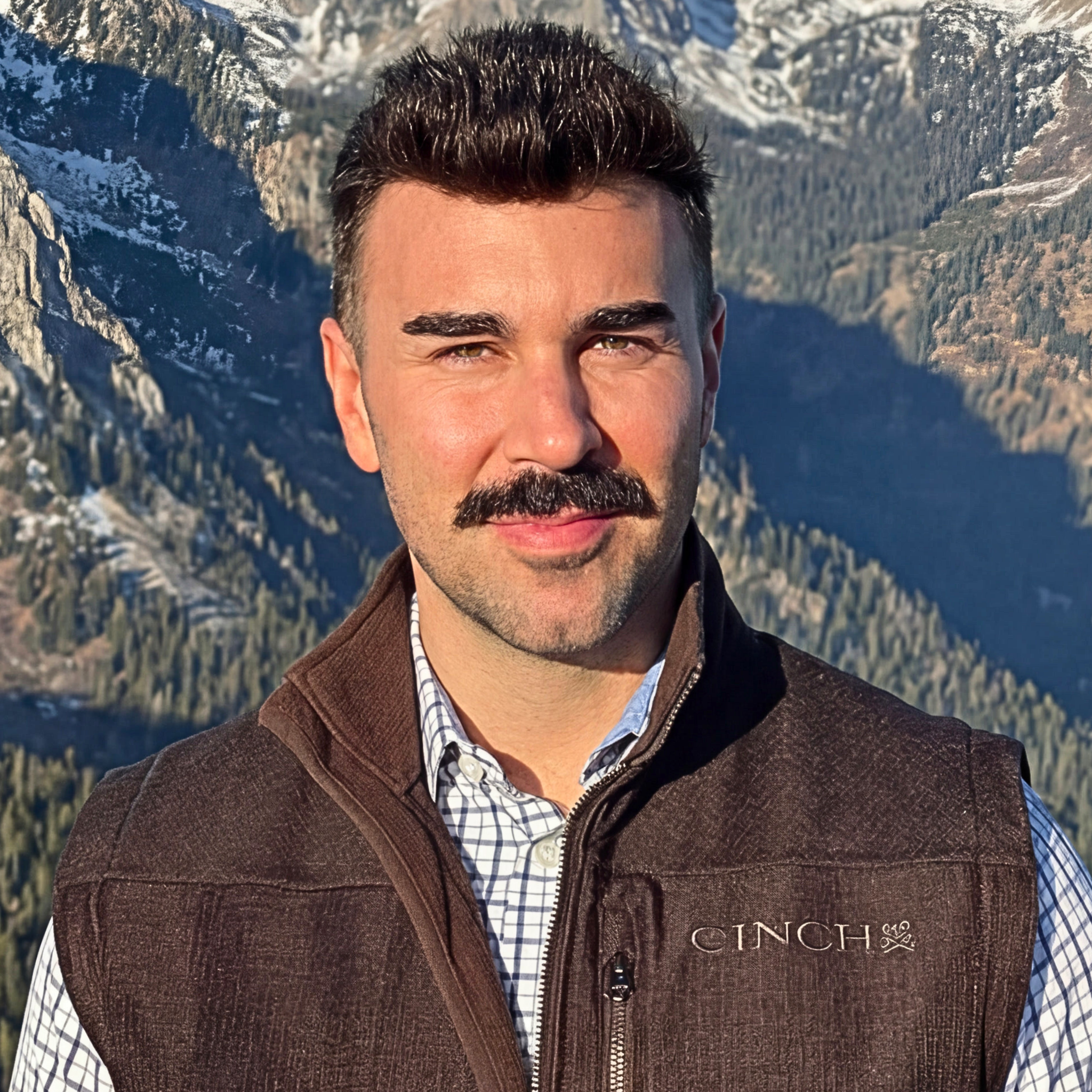 A man with dark hair, a mustache, and light skin, wearing a checkered shirt and a brown vest, standing outdoors with mountains and a lake in the background.