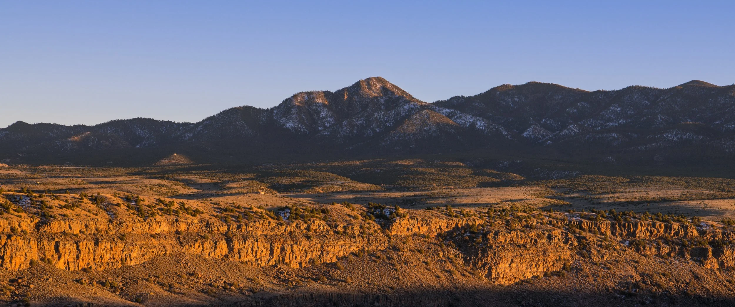Sunset over rugged mountains with snow patches and a dry, rocky foreground with sparse vegetation.