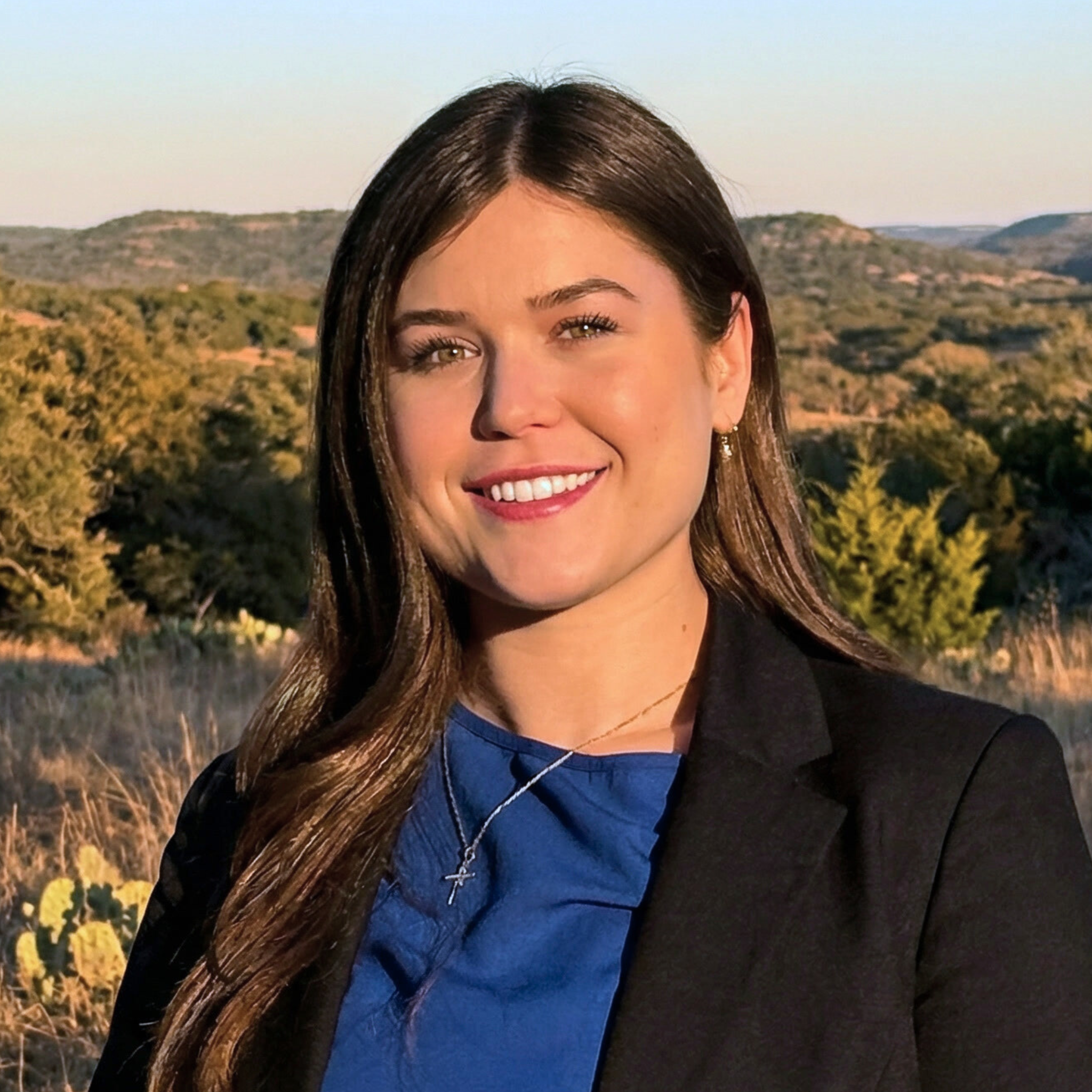 A young woman with long brown hair, smiling, standing outdoors with mountains and greenery in the background during sunset.