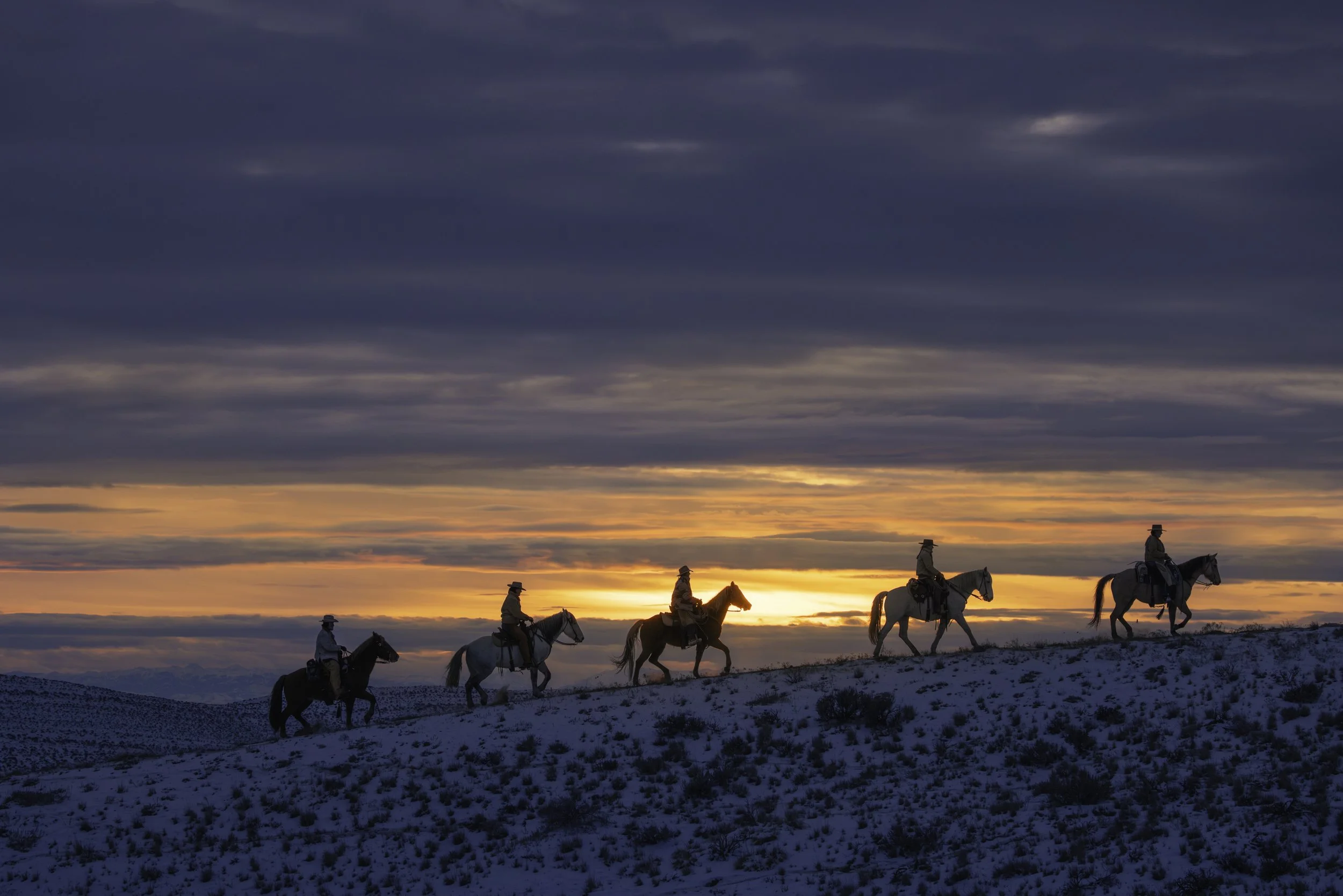 Five people riding horses on a snow-covered hillside during sunset with a cloudy sky.