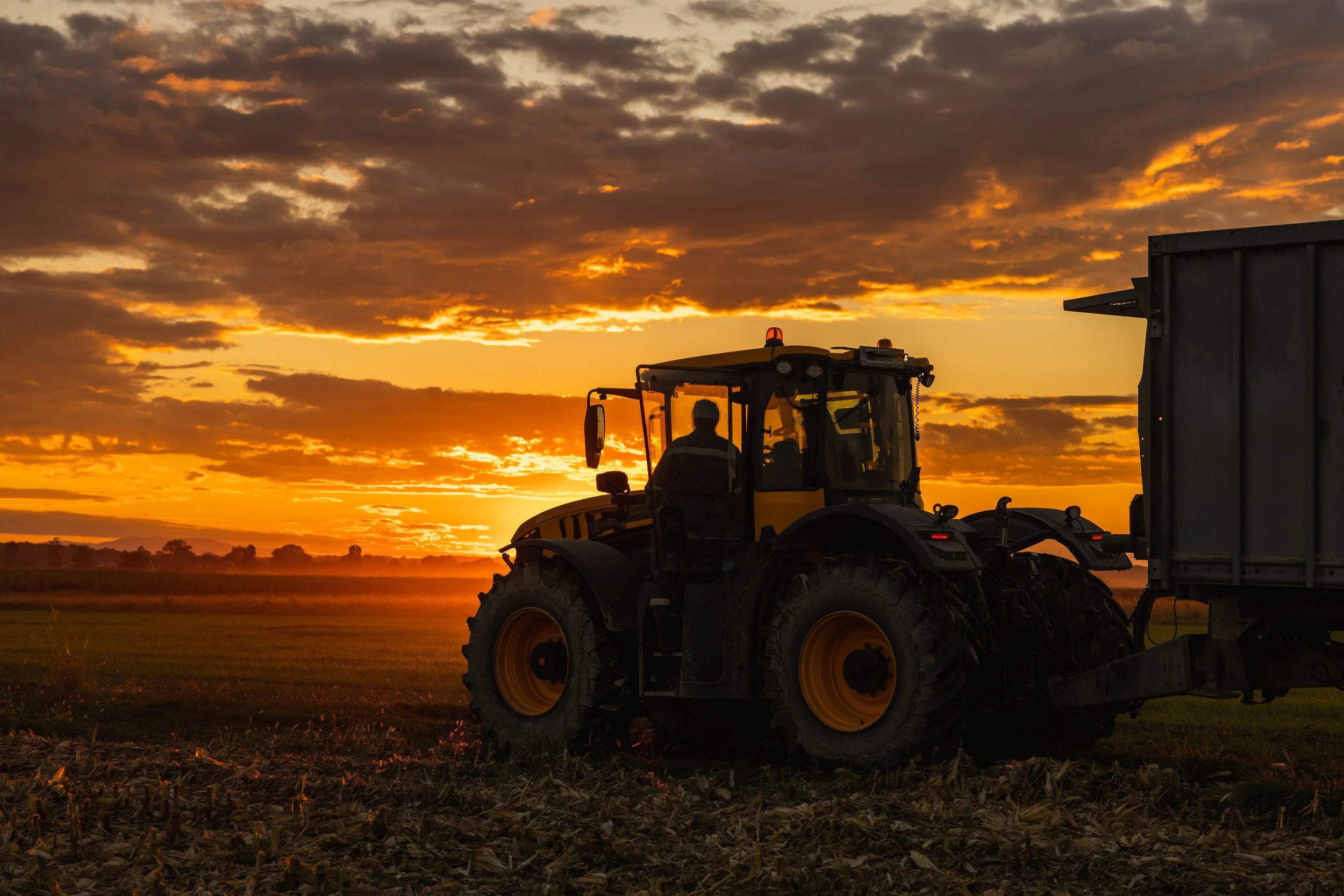 Silhouette of a tractor working in a field at sunset with a cloudy sky and horizon in the background.