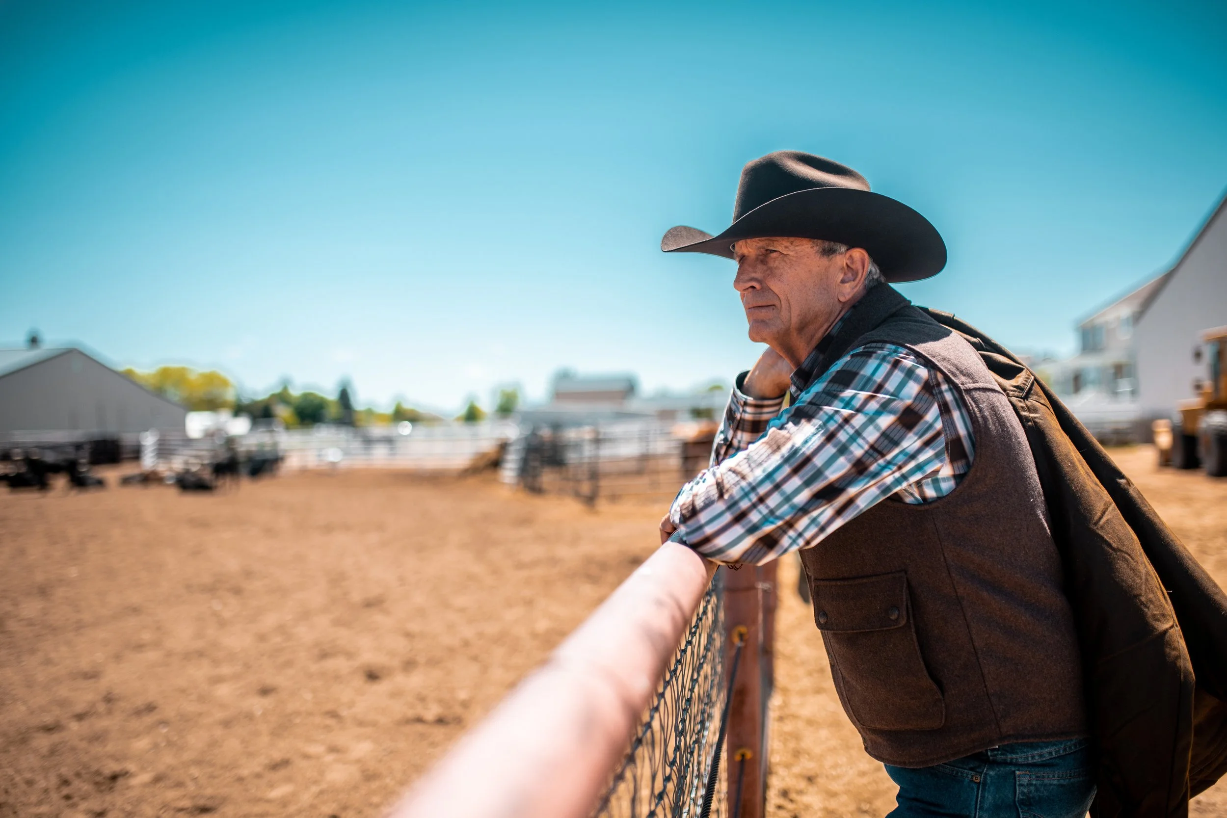 An elderly man wearing a black cowboy hat and plaid shirt leaning on a fence on a farm, looking pensively into the distance.