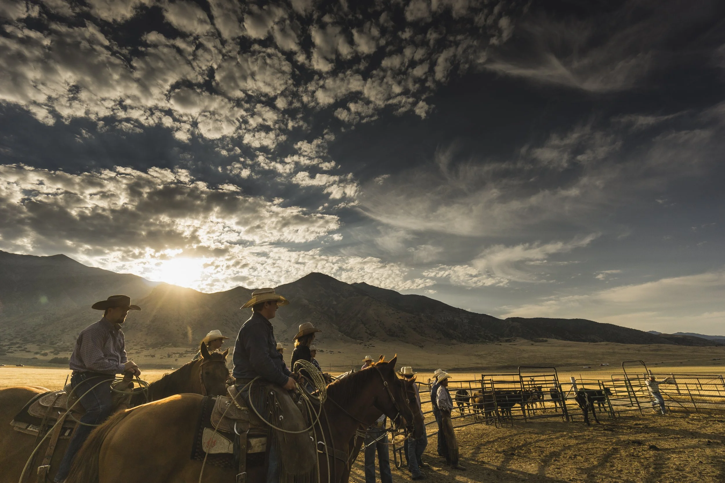 Cowboys on horseback beside cattle pens during sunset on a ranch with mountains and a cloudy sky.