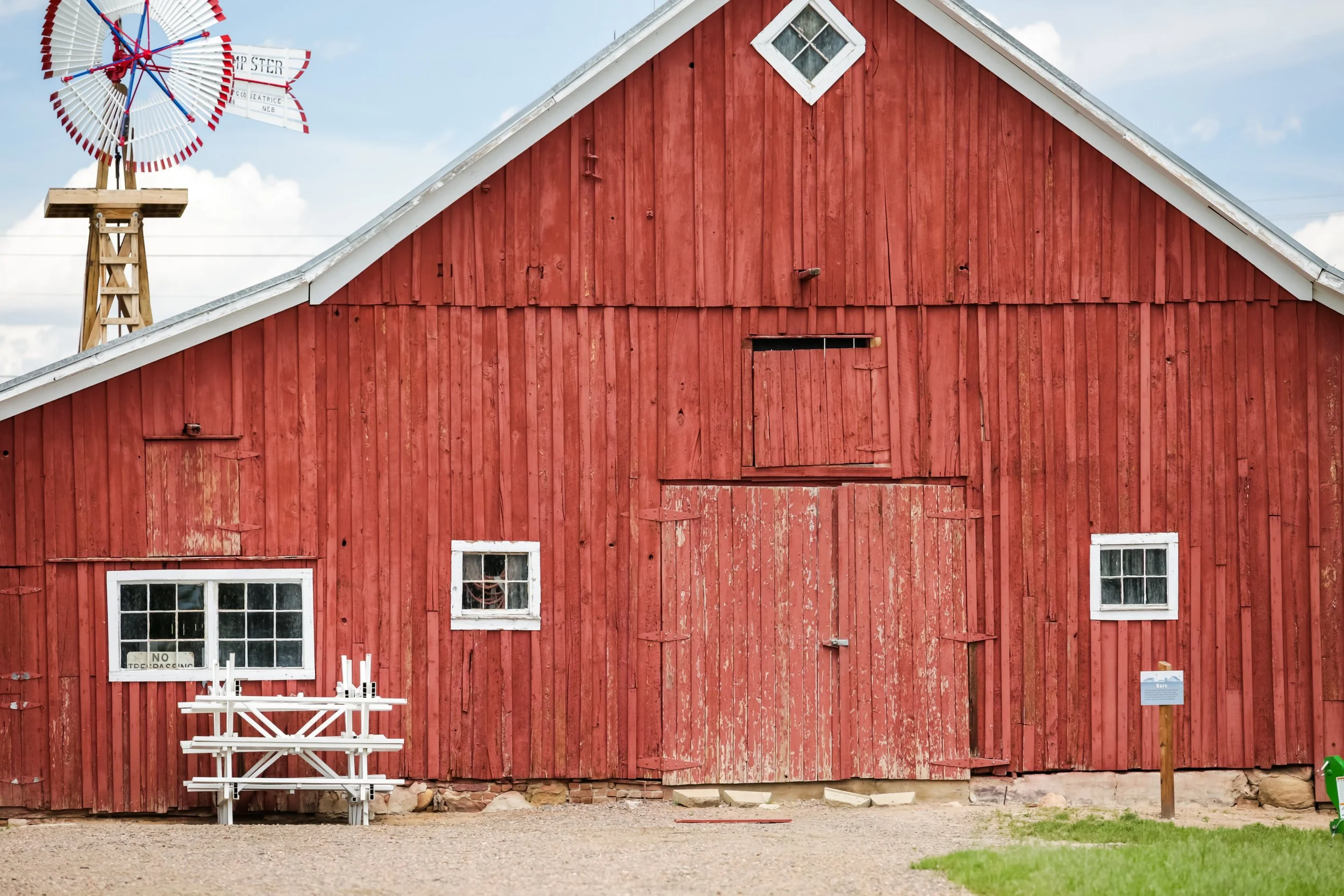 Red barn with white-framed windows, a large door, and a decorative windmill in the background under a partly cloudy sky.