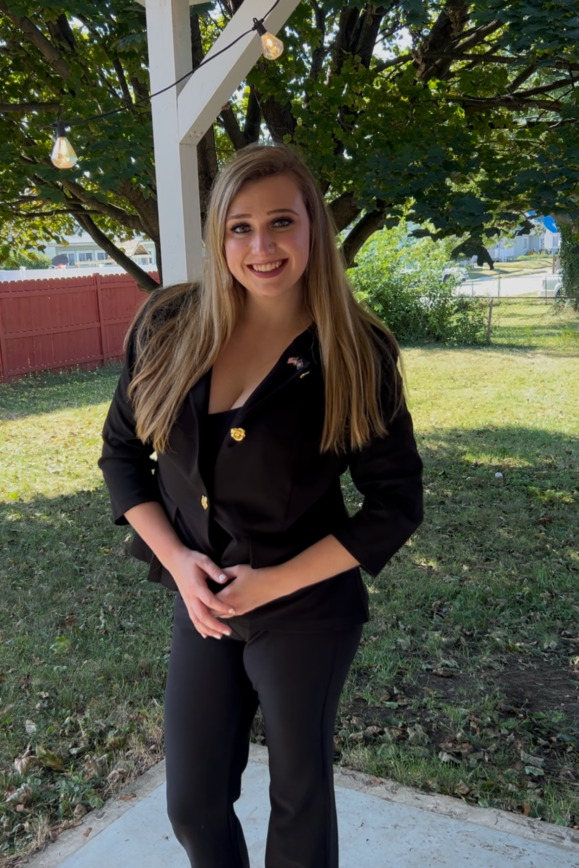 A young woman with long blonde hair smiling outdoors, wearing a black blazer and black pants, standing under a tree with green foliage and string lights hanging above.