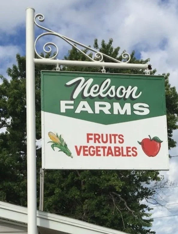 Sign for Nelson Farms advertising fruits and vegetables, with illustrations of a cob of corn, a green pepper, and a red apple.