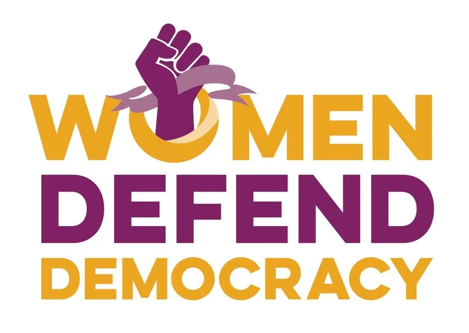 A graphic with the text 'Women Defend Democracy' in bold yellow and purple letters, featuring a purple fist breaking through a yellow ring.