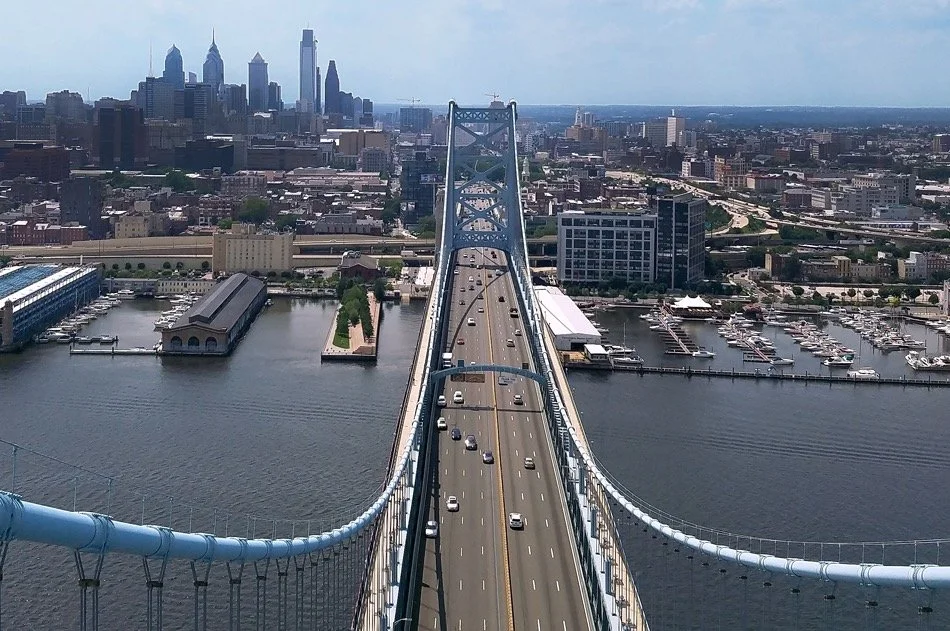 Aerial view of a suspension bridge over a body of water leading to downtown Philadelphia with skyscrapers in the background.
