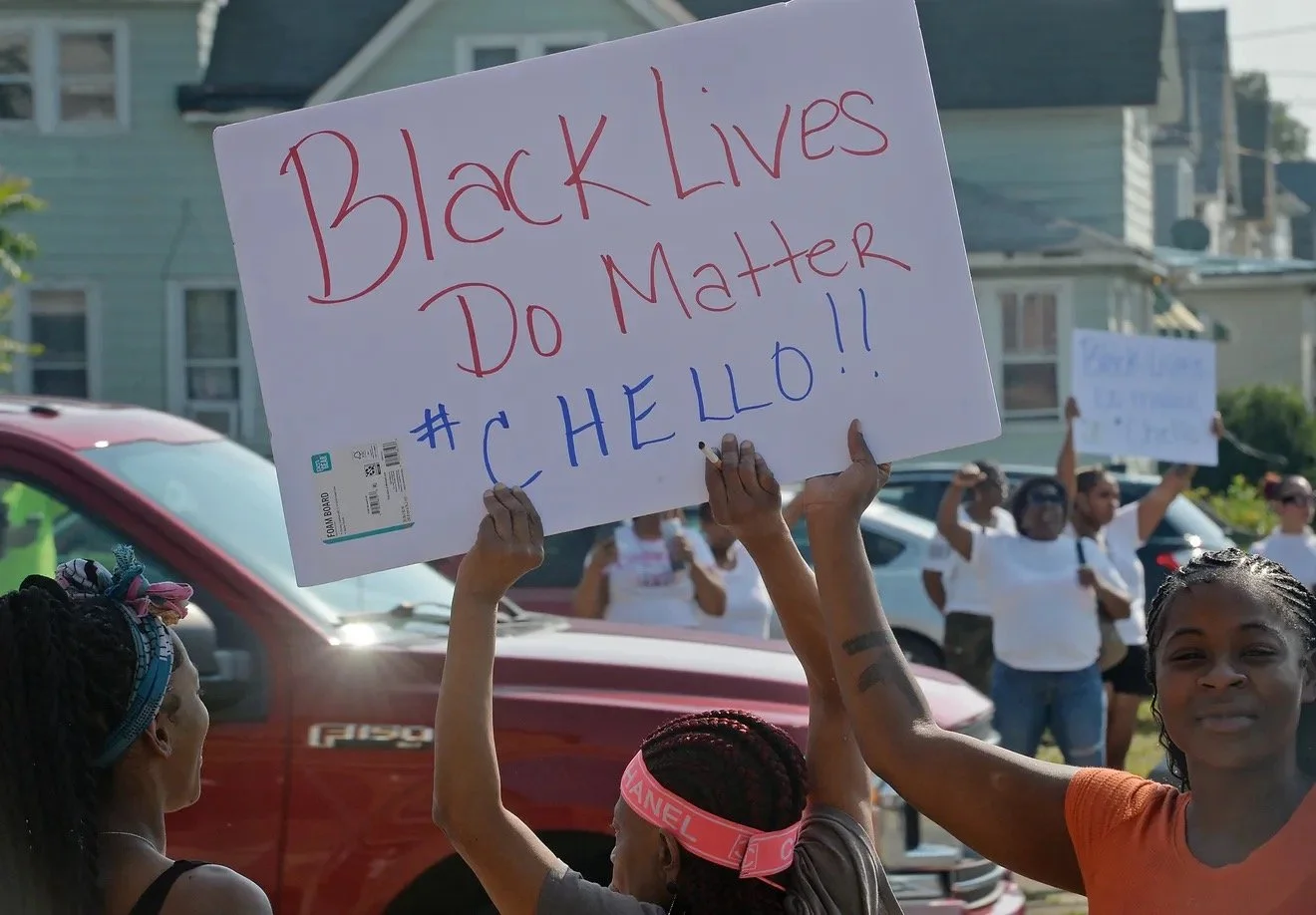 Protesters holding signs during a Black Lives Matter demonstration, with one sign reading 'Black Lives Do Matter #HELLO!!' in an urban neighborhood.