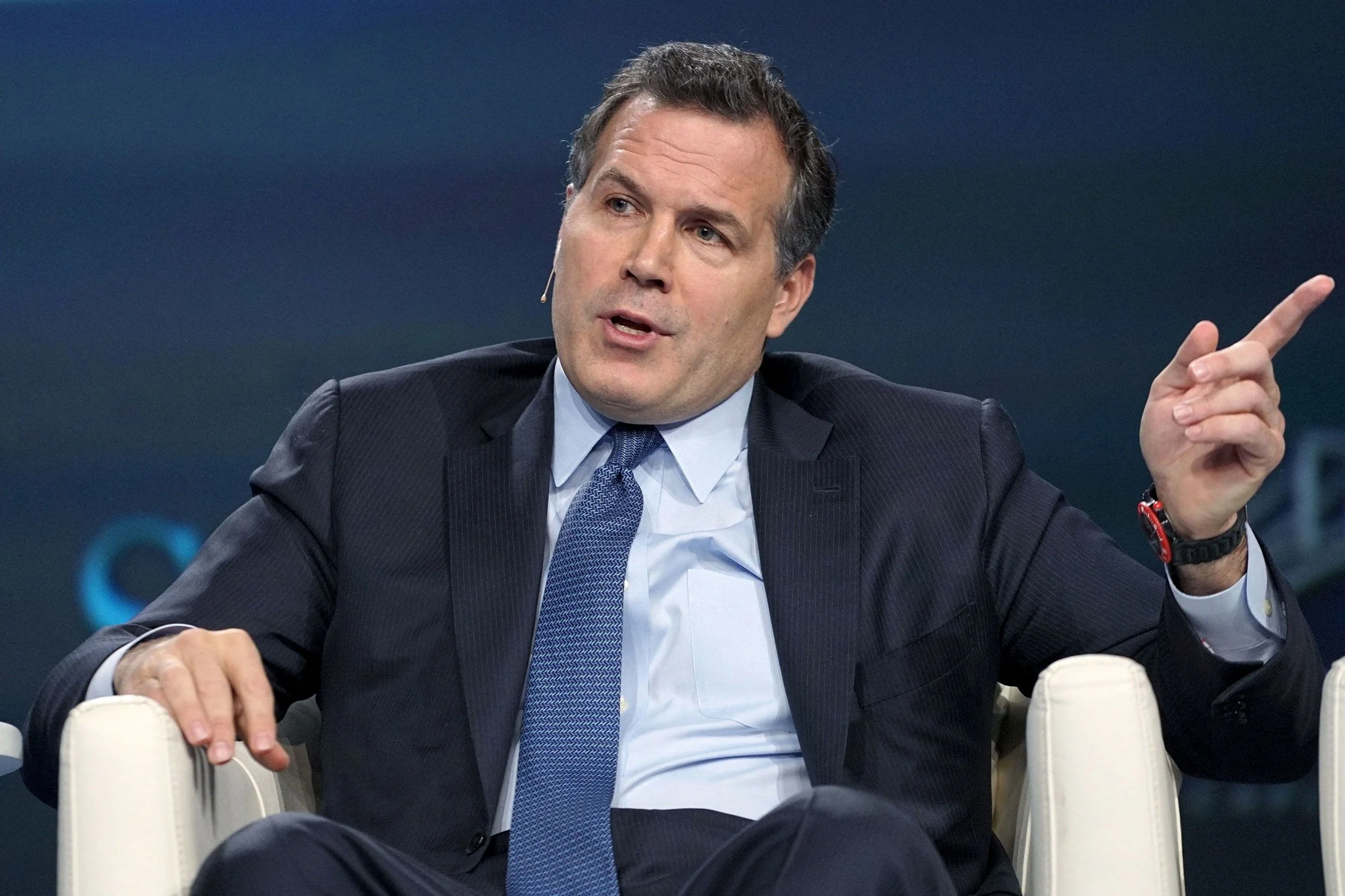 A man in a dark suit and blue tie, speaking or making a point, sitting on a white chair with a dark background.