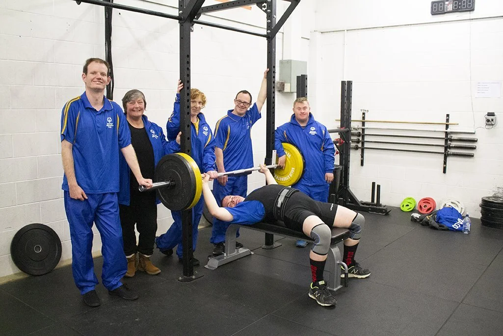 A young woman with a black workout outfit and knee wraps bench presses with a barbell in a gym, surrounded by five smiling people in matching blue uniforms, some holding the barbell and others standing nearby. There are gym equipment and weight plates visible in the background.