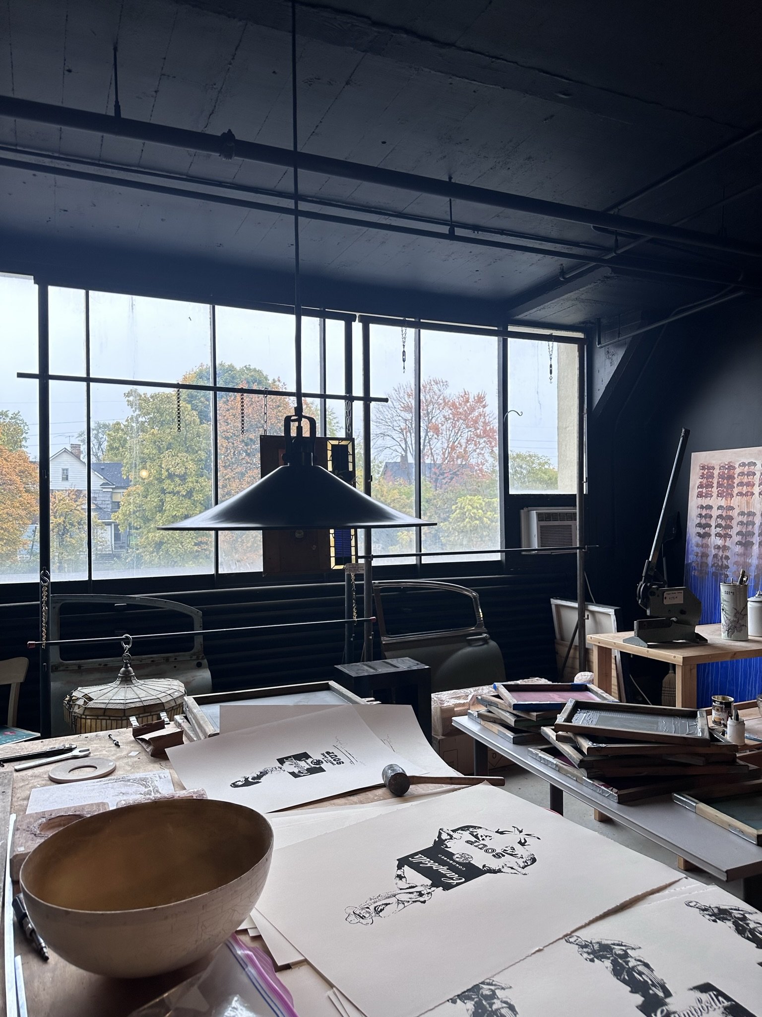 Art studio workspace with sketches, books, and art supplies on tables, large window with autumn trees outside, and studio equipment like a lamp and an air conditioner.
