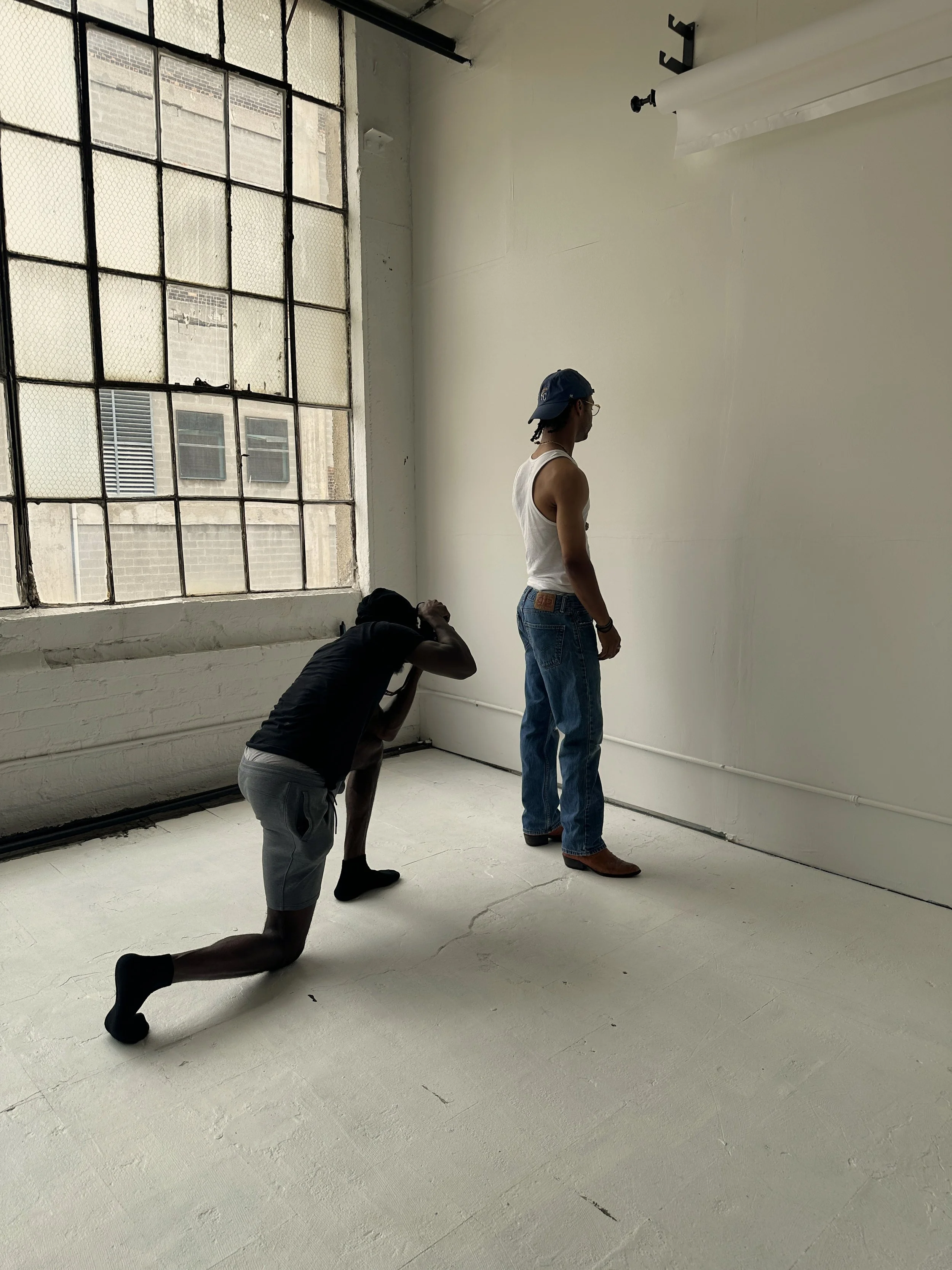 Two people are in an art studio with large industrial windows. One person is in a kneeling pose, trying to mimic the other person who is standing and facing the wall, with a plain wall as background.