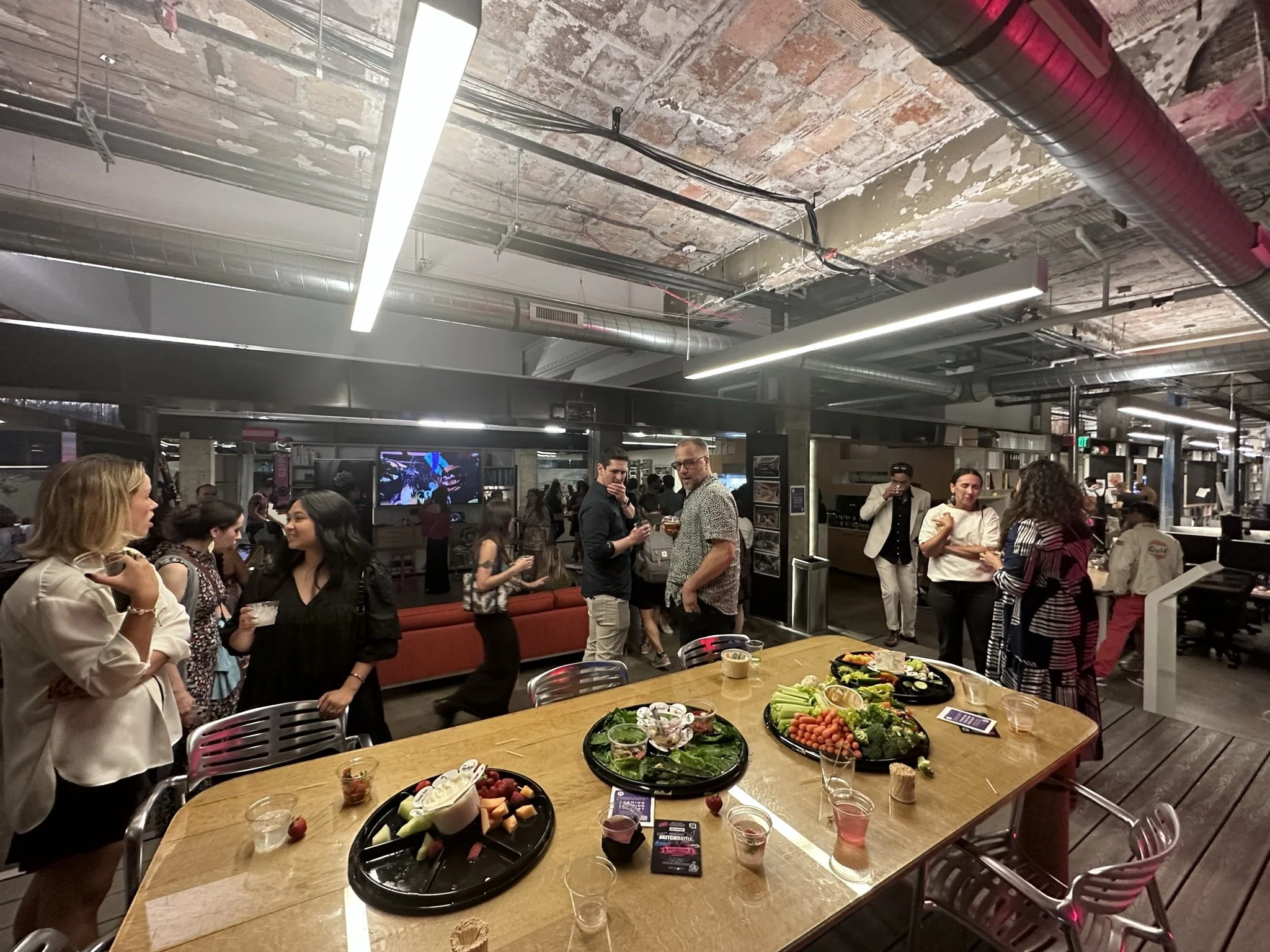 People socializing at a bar or restaurant with food and drinks on a large wooden table, in an industrial-style space with exposed brick ceiling.