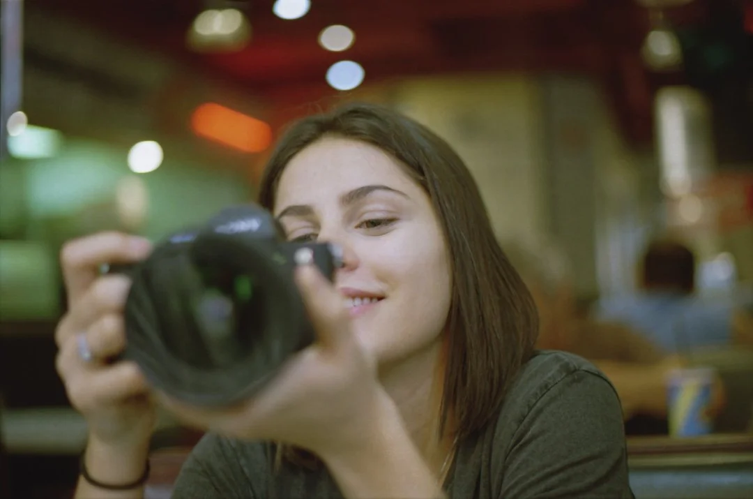 A woman with dark brown hair smiling as she looks through the viewfinder of a camera in a colorful, well-lit indoor space, possibly a diner or cafe.