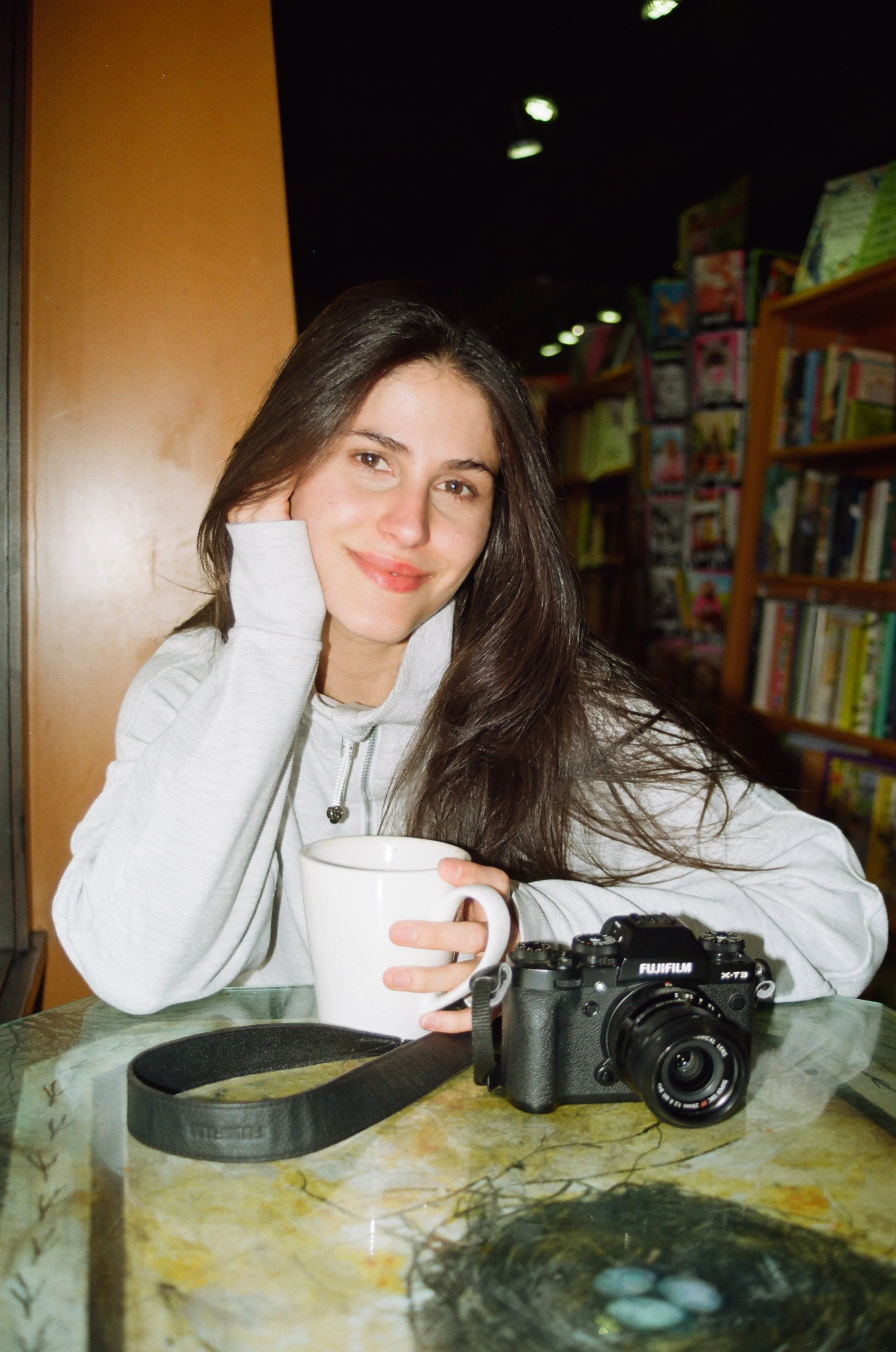 A young woman with long brown hair sitting at a marble table in a bookstore, holding a white mug, with a camera on the table and bookshelves in the background.