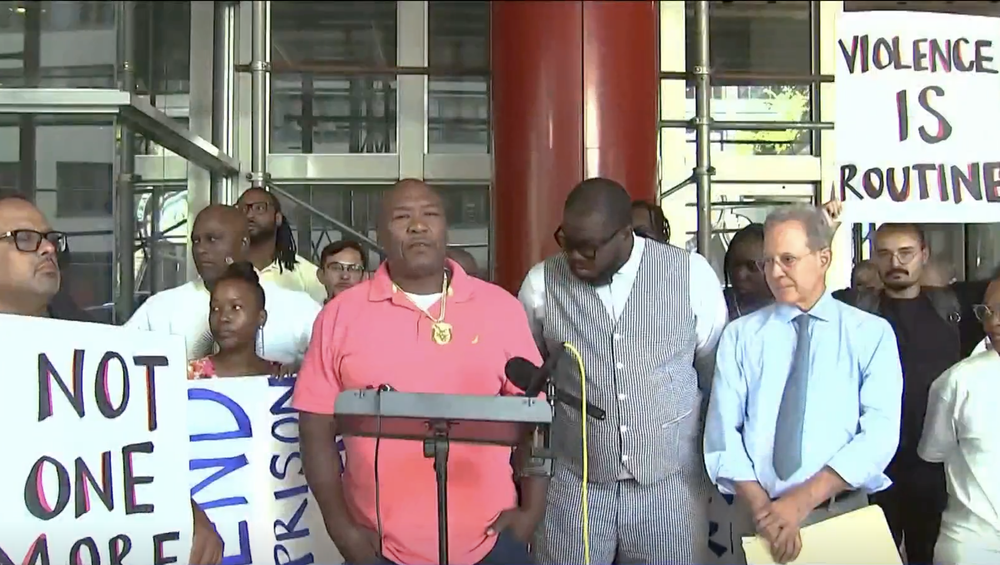 Man at podium surrounded by crowd with signs reading End Prison Violence
