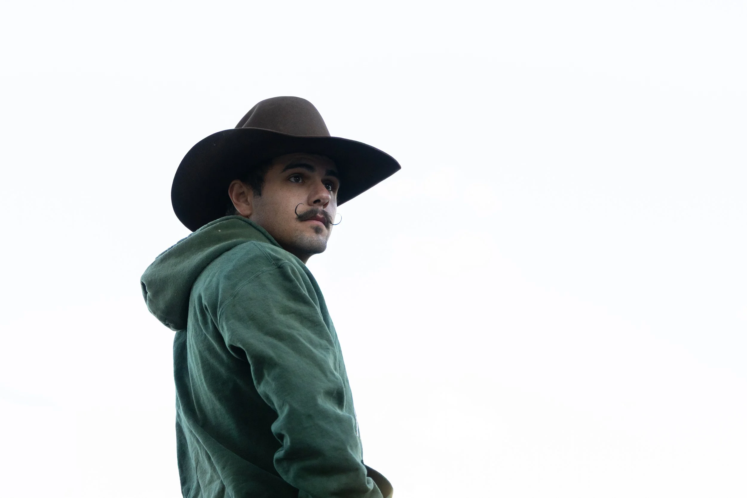 A man wearing a large cowboy hat and a green jacket, with a well-groomed mustache, looking off to the side against a cloudy sky.