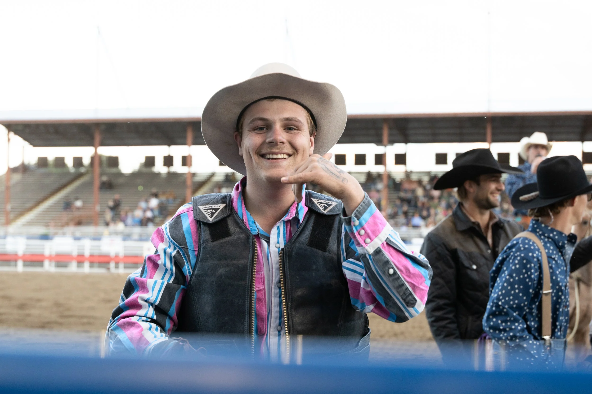 A smiling young man wearing a cowboy hat, colorful striped shirt, and safety vest standing at a rodeo or equestrian event with spectators and other participants in cowboy hats in the background.