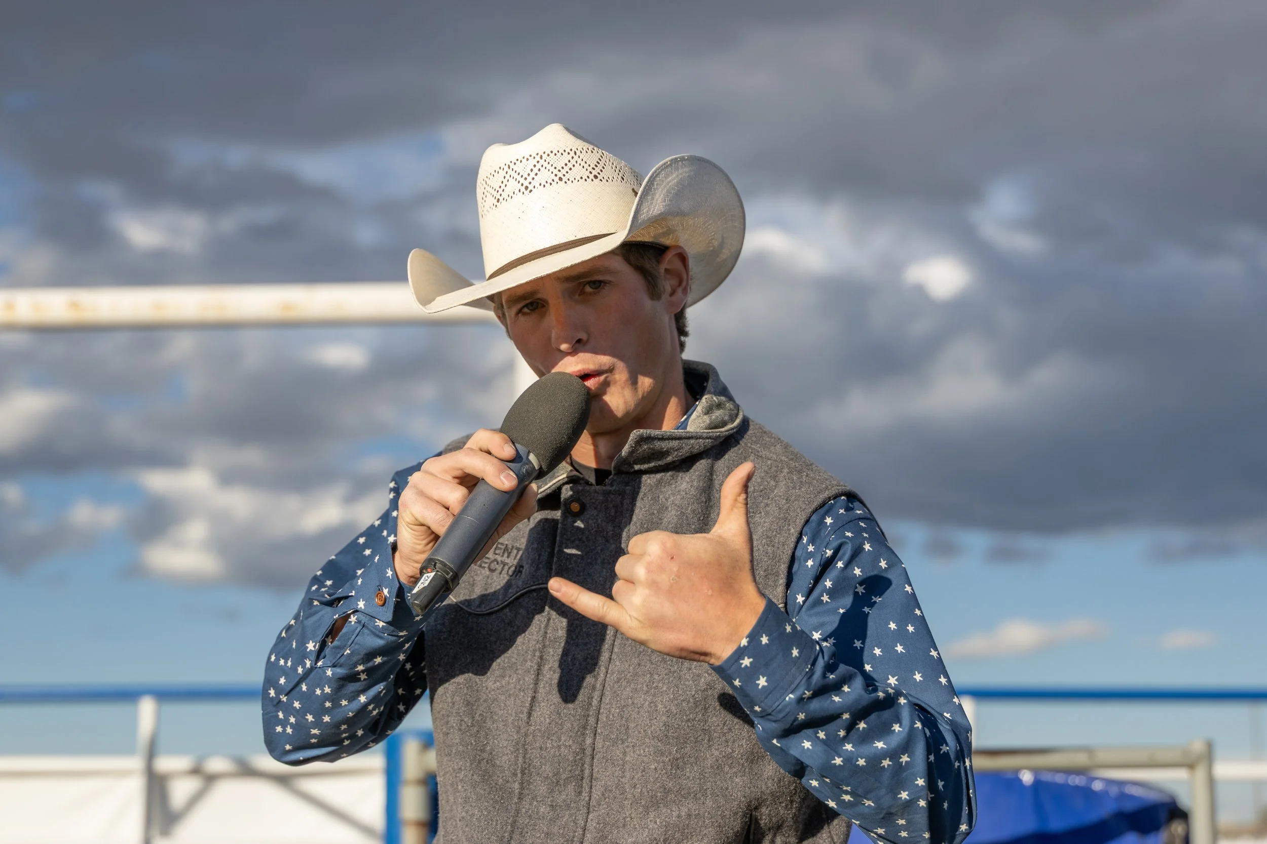 Young man wearing a white cowboy hat, a gray vest with a patch on it, and a blue shirt with white stars, holding a microphone, outdoors on a cloudy day.