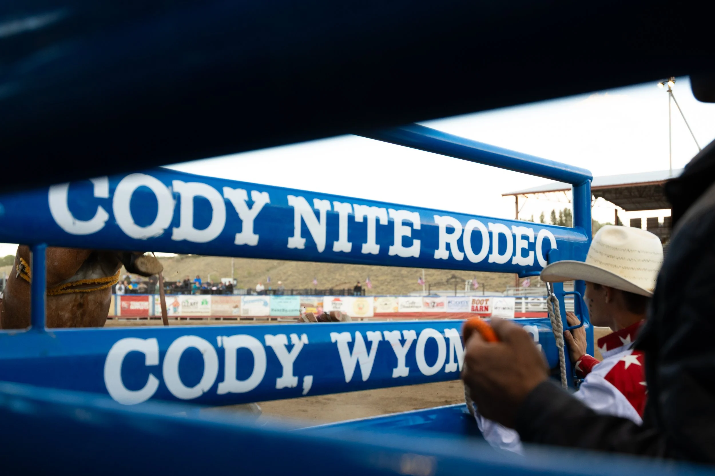 Close-up of a rodeo float with blue railing displaying white text, at an outdoor rodeo arena with spectators and participants in Western attire, including cowboy hats and boots.