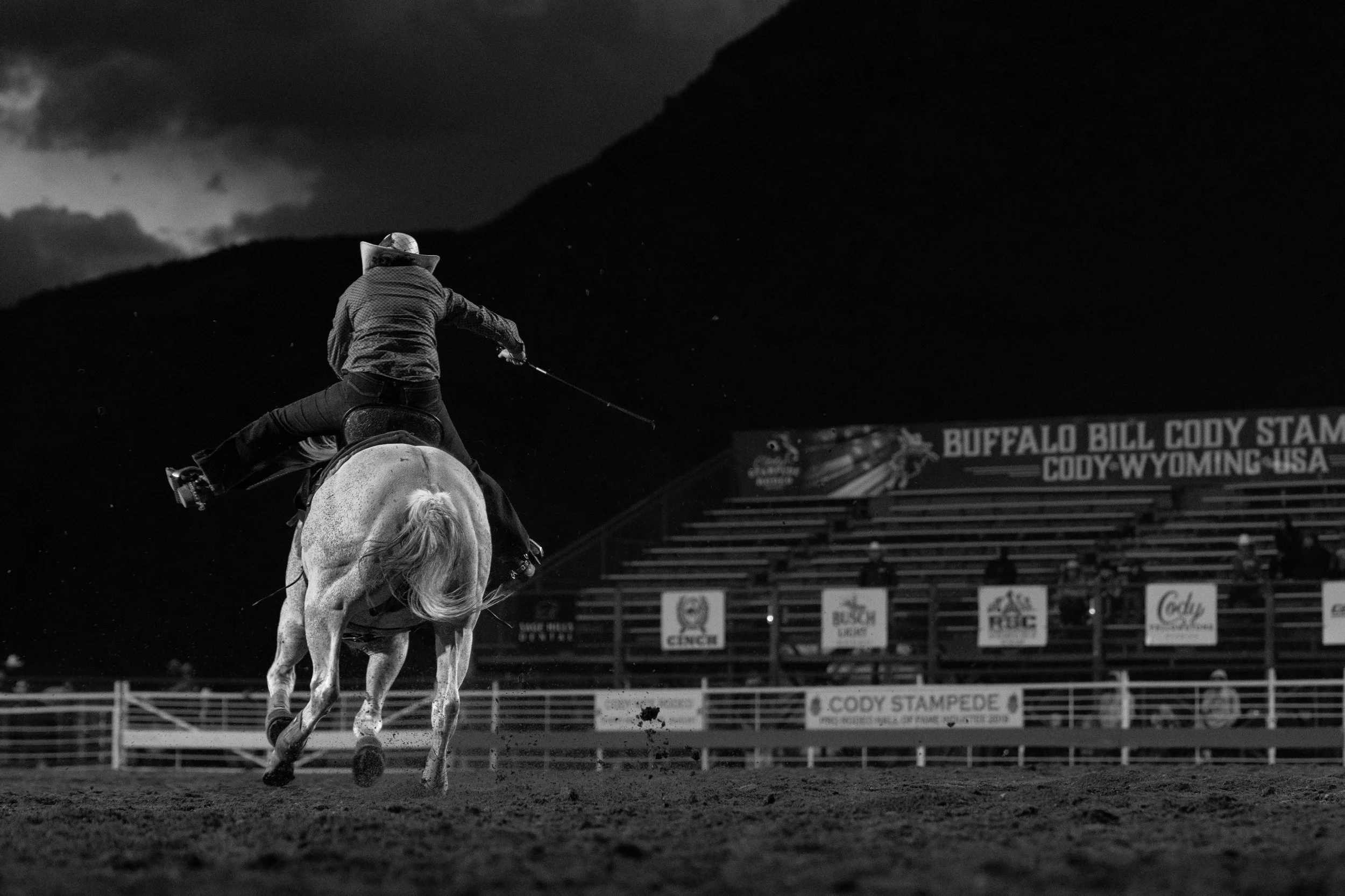 Cowboy riding a bucking horse during a rodeo event at Cody Stampede in Cody, Wyoming, USA. The cowboy wears a hat and western attire, with a backdrop of empty bleachers and a cloudy sky.