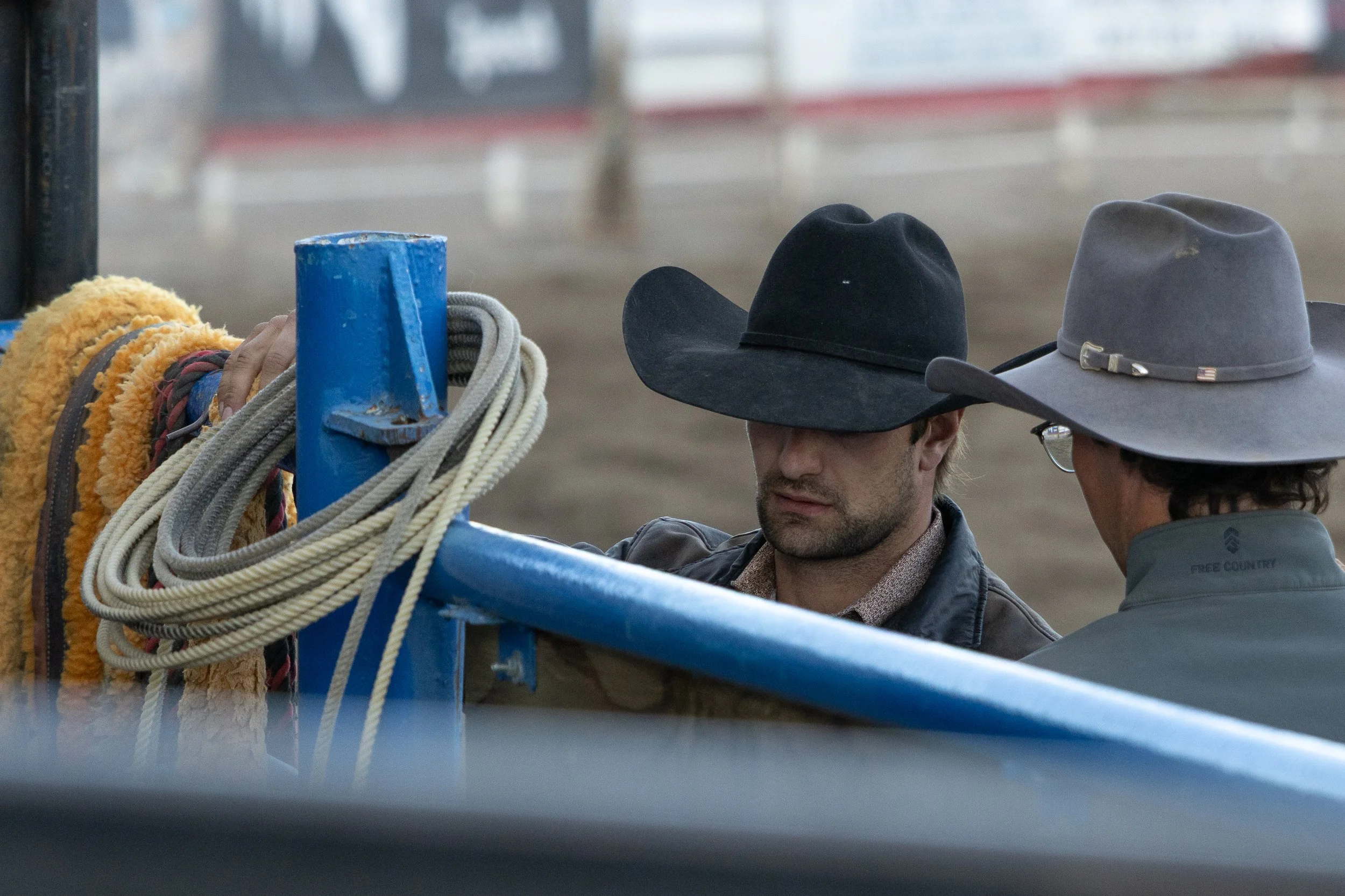 Two men wearing cowboy hats and jackets are near a blue metal structure with ropes and equipment hanging on it, in a possibly outdoor or rodeo setting.