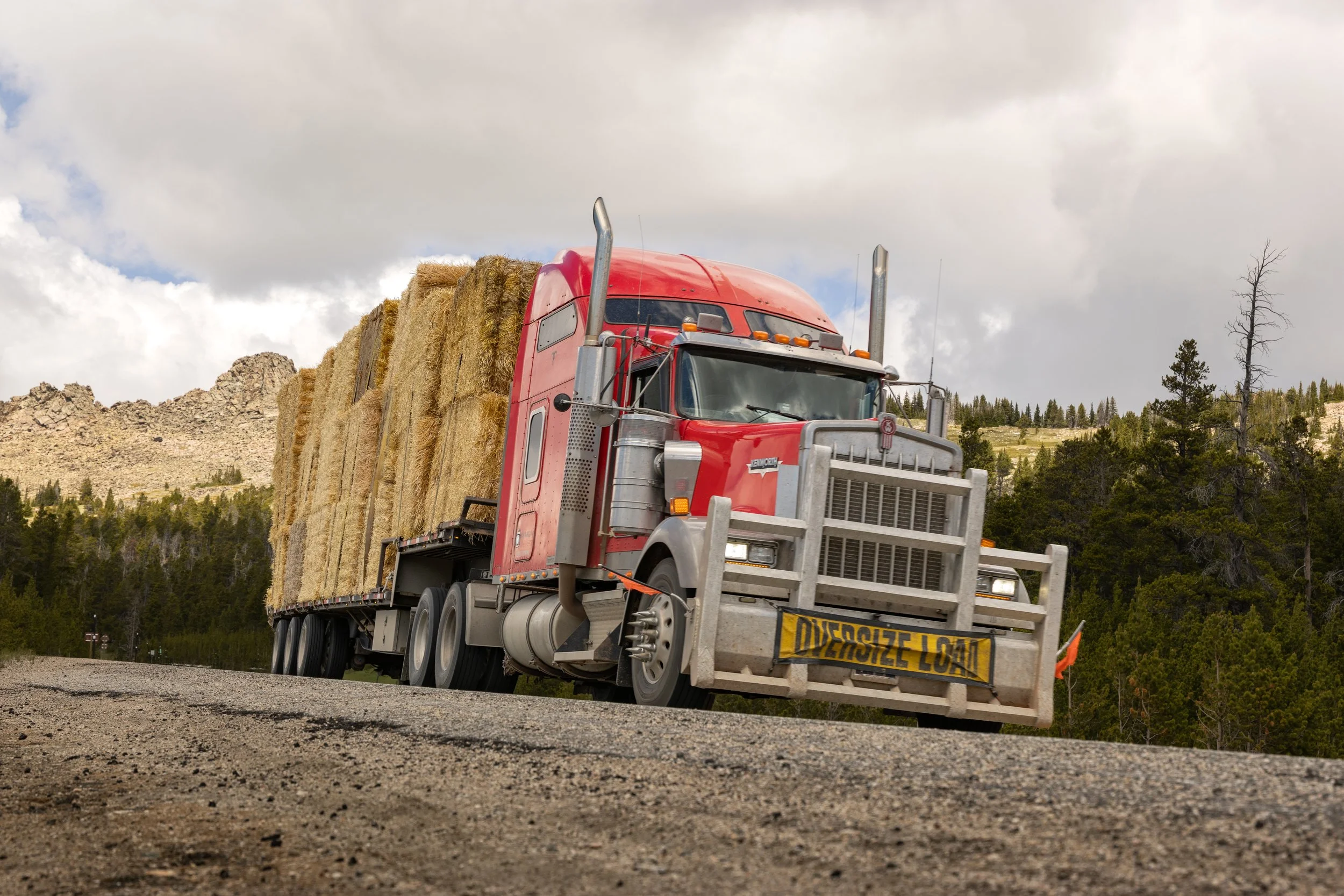 A red semi-truck carrying hay bales on a highway with a mountainous landscape and cloudy sky in the background.