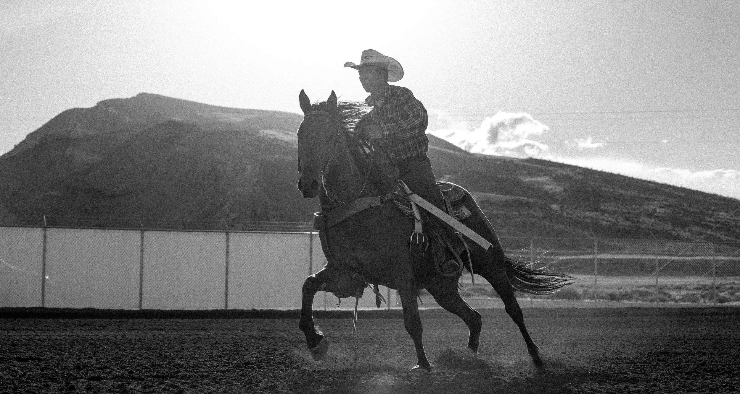 A person riding a horse in an outdoor arena, with a mountain in the background, all in black and white.