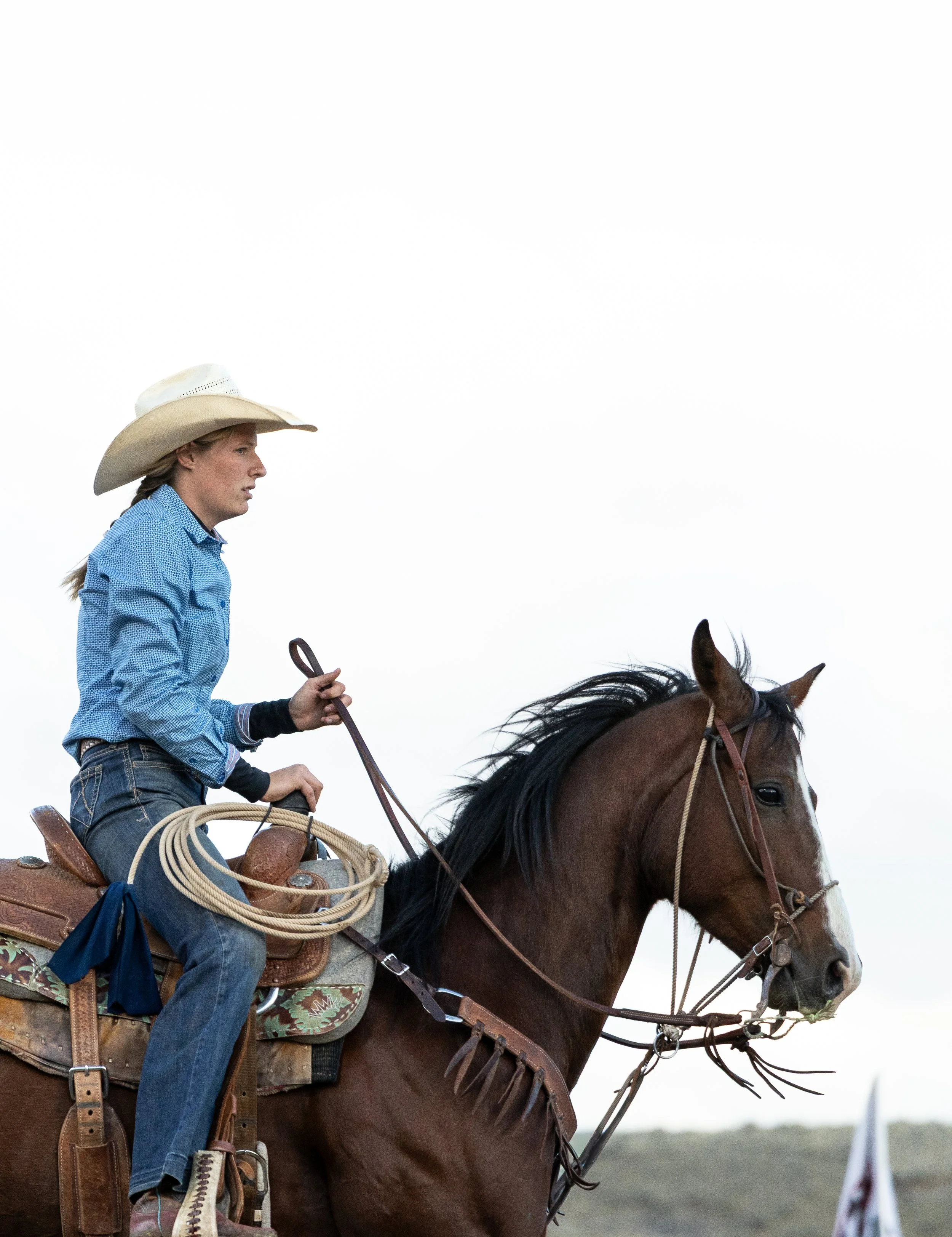 A woman wearing a cowboy hat and blue shirt riding a brown horse with a saddle and reins in an open plain with a cloudy sky.