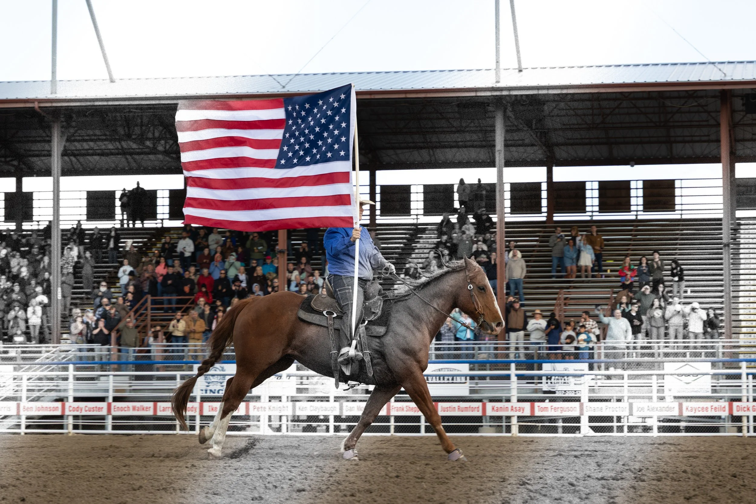 A person riding a horse in a rodeo arena, holding an American flag, with spectators in the stands behind.