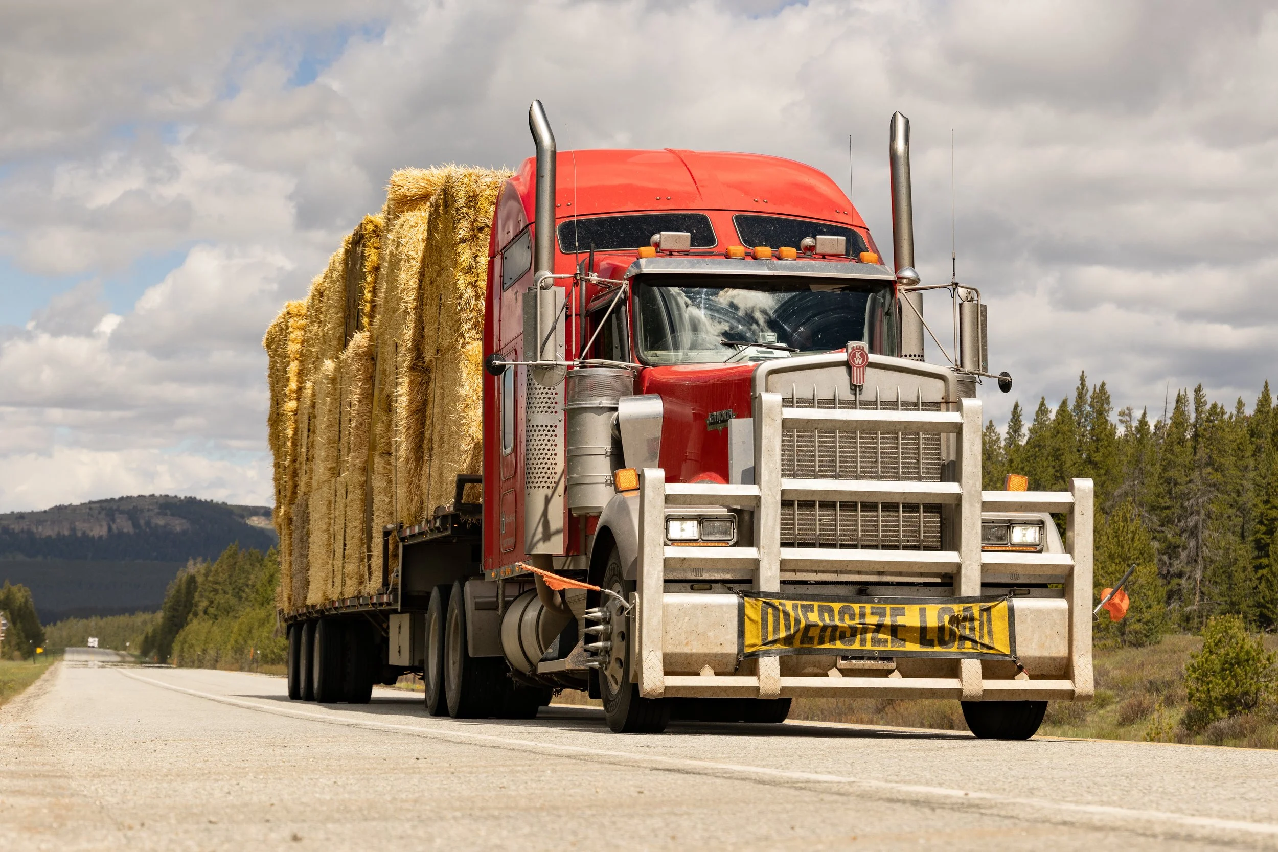 A red semi-truck carrying hay bales on a road with a cloudy sky and forested hills in the background.
