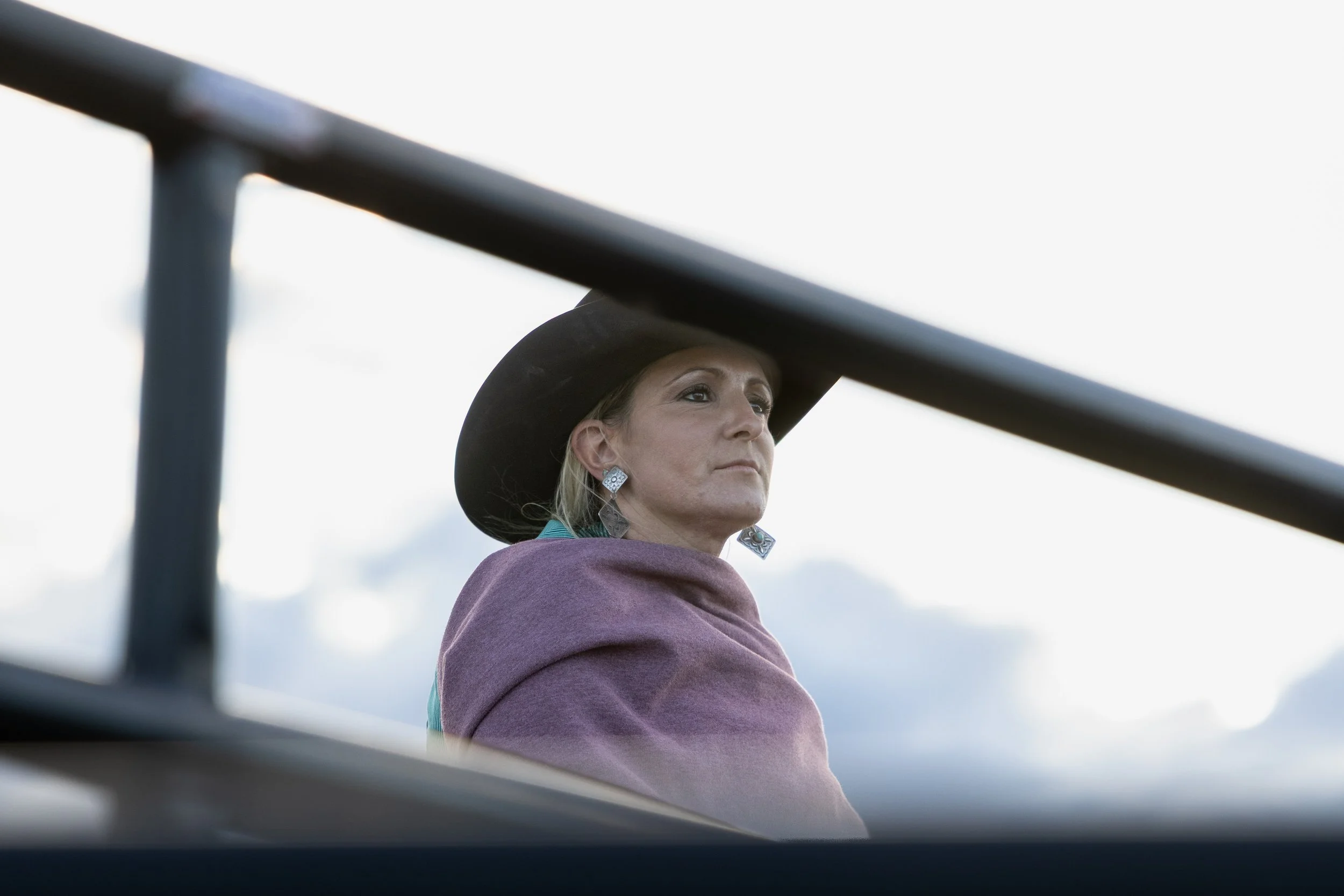 A woman wearing a wide-brimmed black hat and diamond earrings, looking into the distance with a serious expression, seen from a low angle through a metal railing.